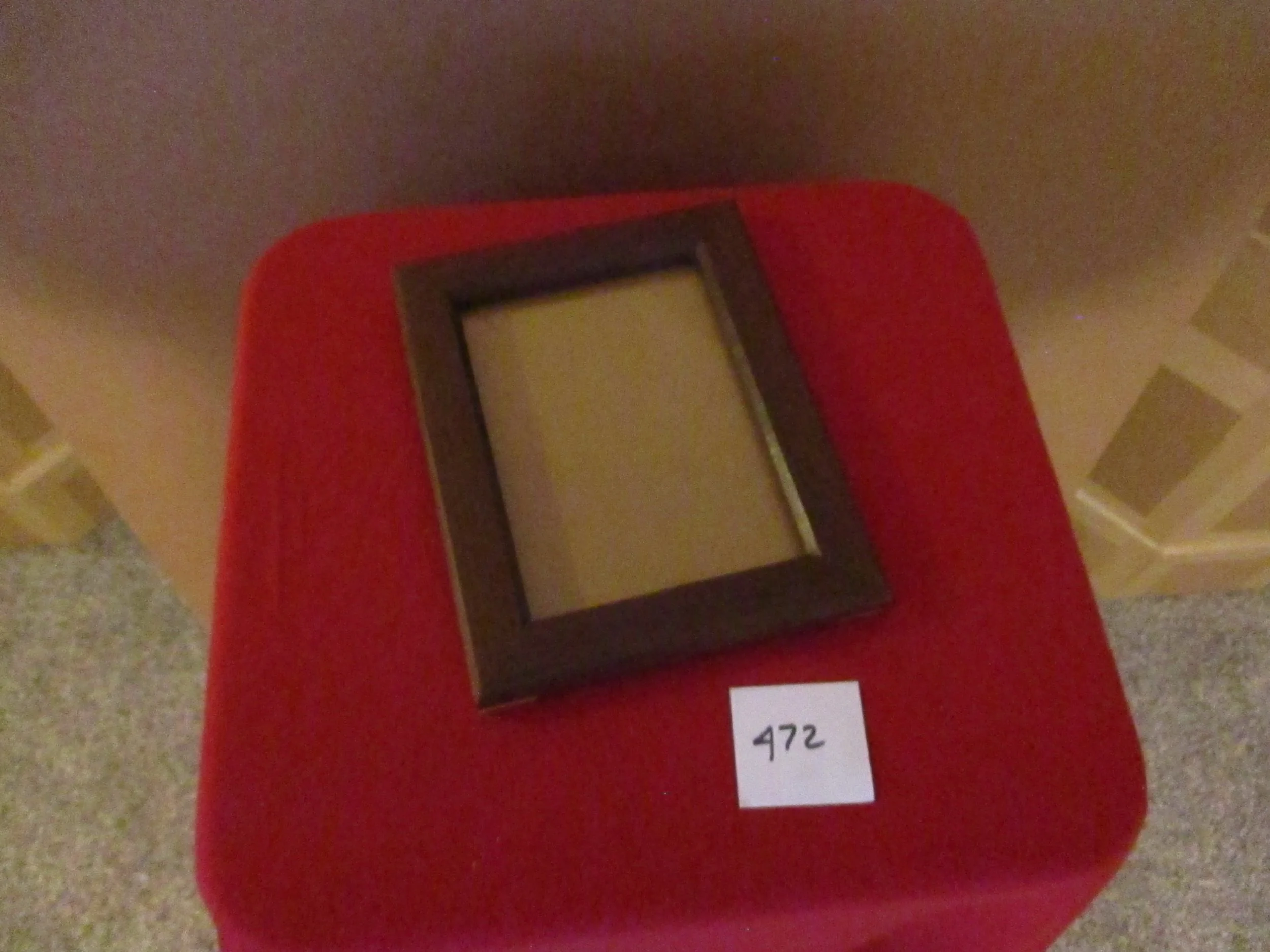 A wooden picture frame with a dark brown border resting on a red cushioned surface. There is a white label with the number 472 placed below the frame.