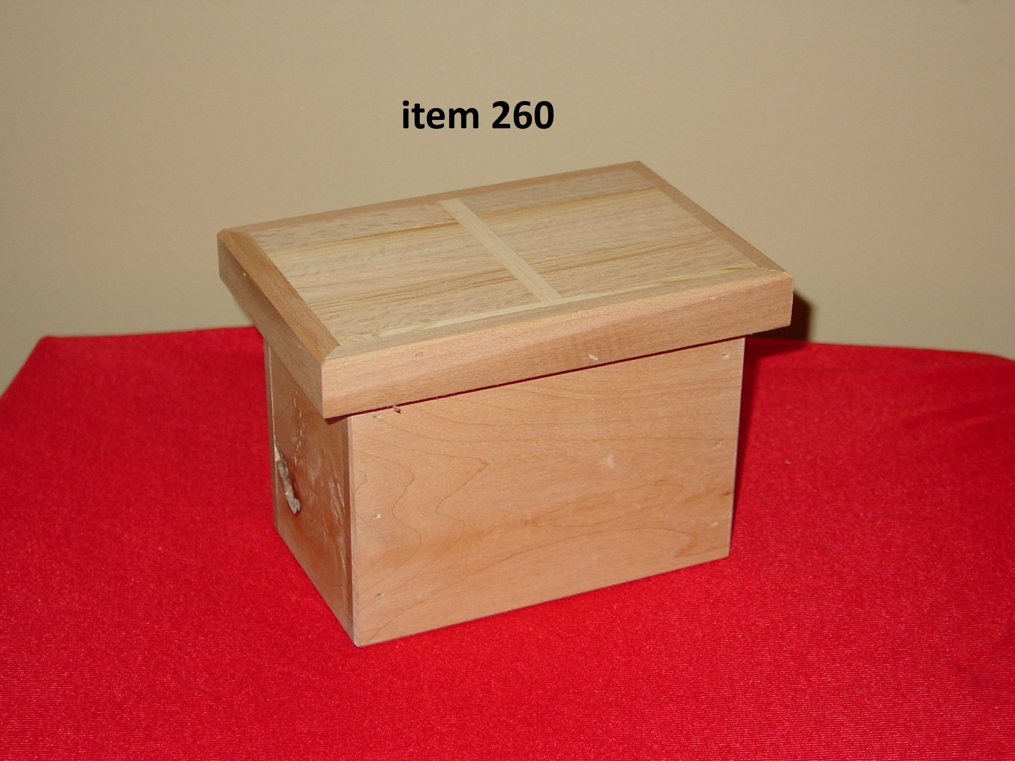 A wooden box with a slanted lid, placed on a red surface against a beige wall. The box has a natural wood finish and appears handmade.