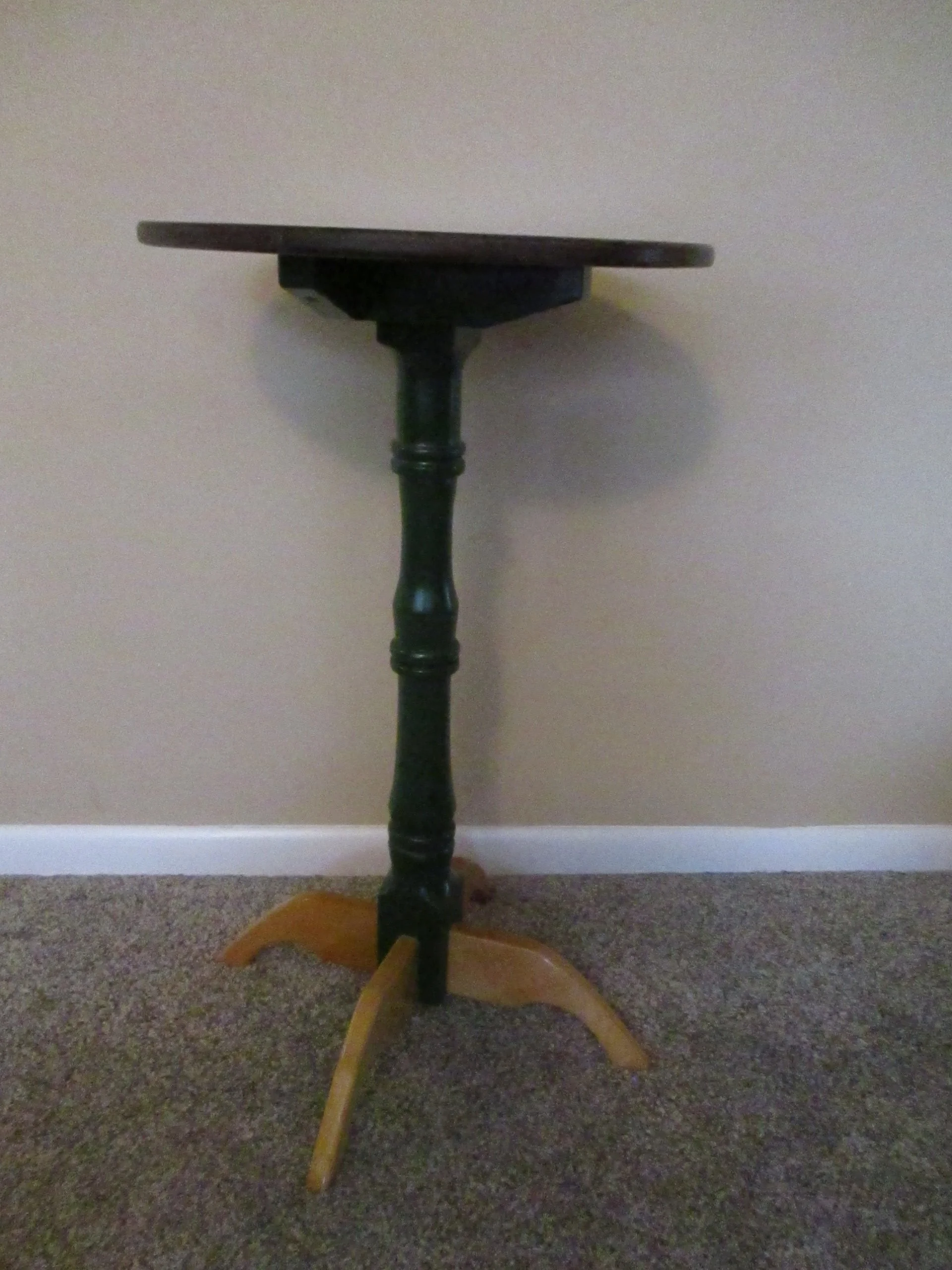Old-fashioned wooden side table with a round top, turned leg, and three curved legs on carpeted floor against a beige wall.
