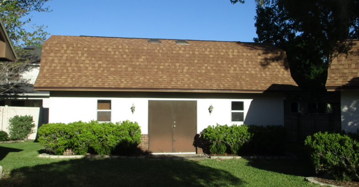 Front view of a house with a brown roof, white walls, and a large metal door, surrounded by green bushes and a lawn, under a clear blue sky.