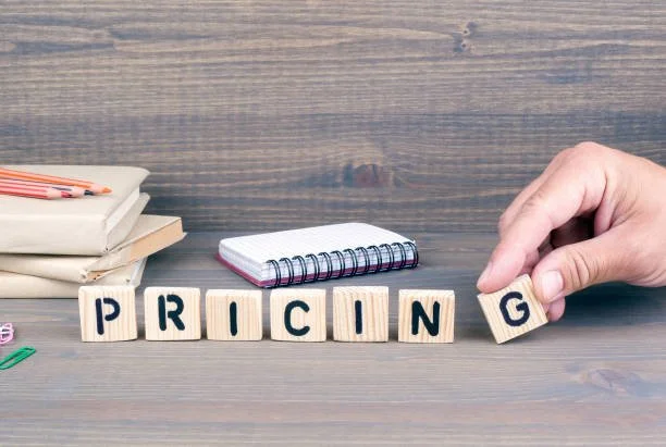 A hand placing a wooden block spell for "PRICING" on a wooden table, with notebooks and pencils in the background.