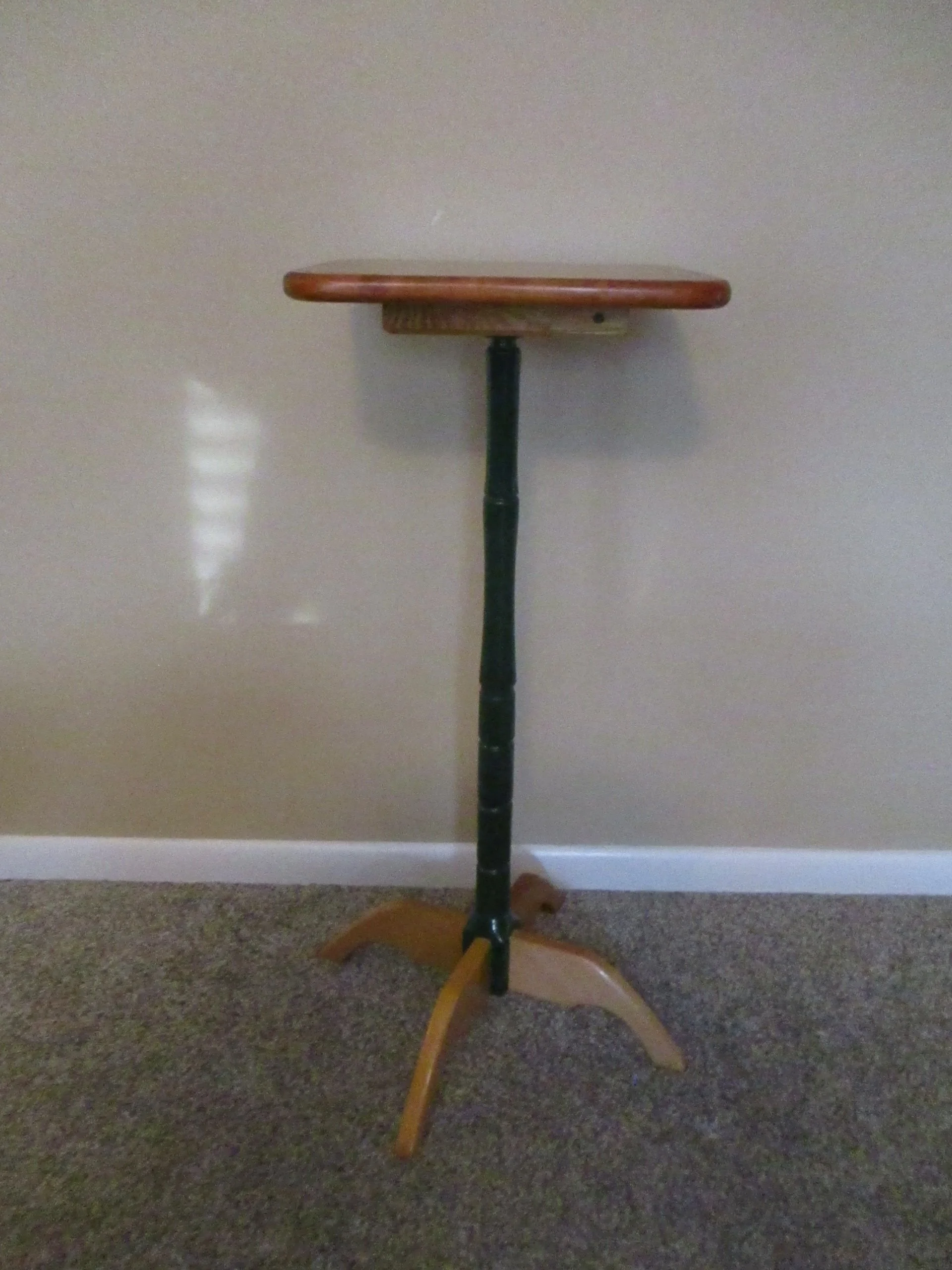 An empty wooden and metal standing pedestal table with a round top, standing against a plain beige wall on a carpeted floor.