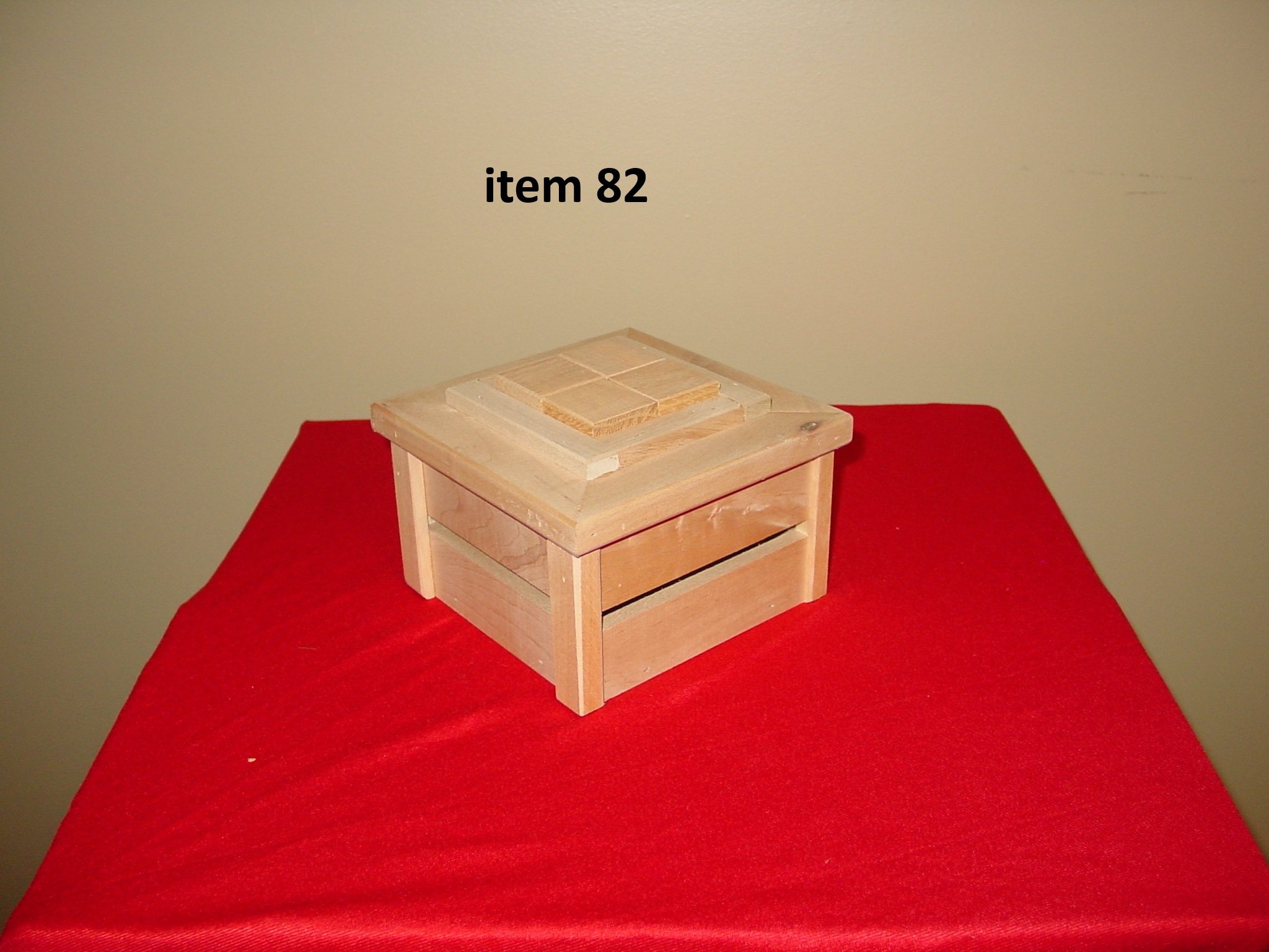 A small wooden box with a slatted drawer on a red cloth-covered surface, against a plain wall. The top of the box has a tiled design with the text 'item 82' above it.