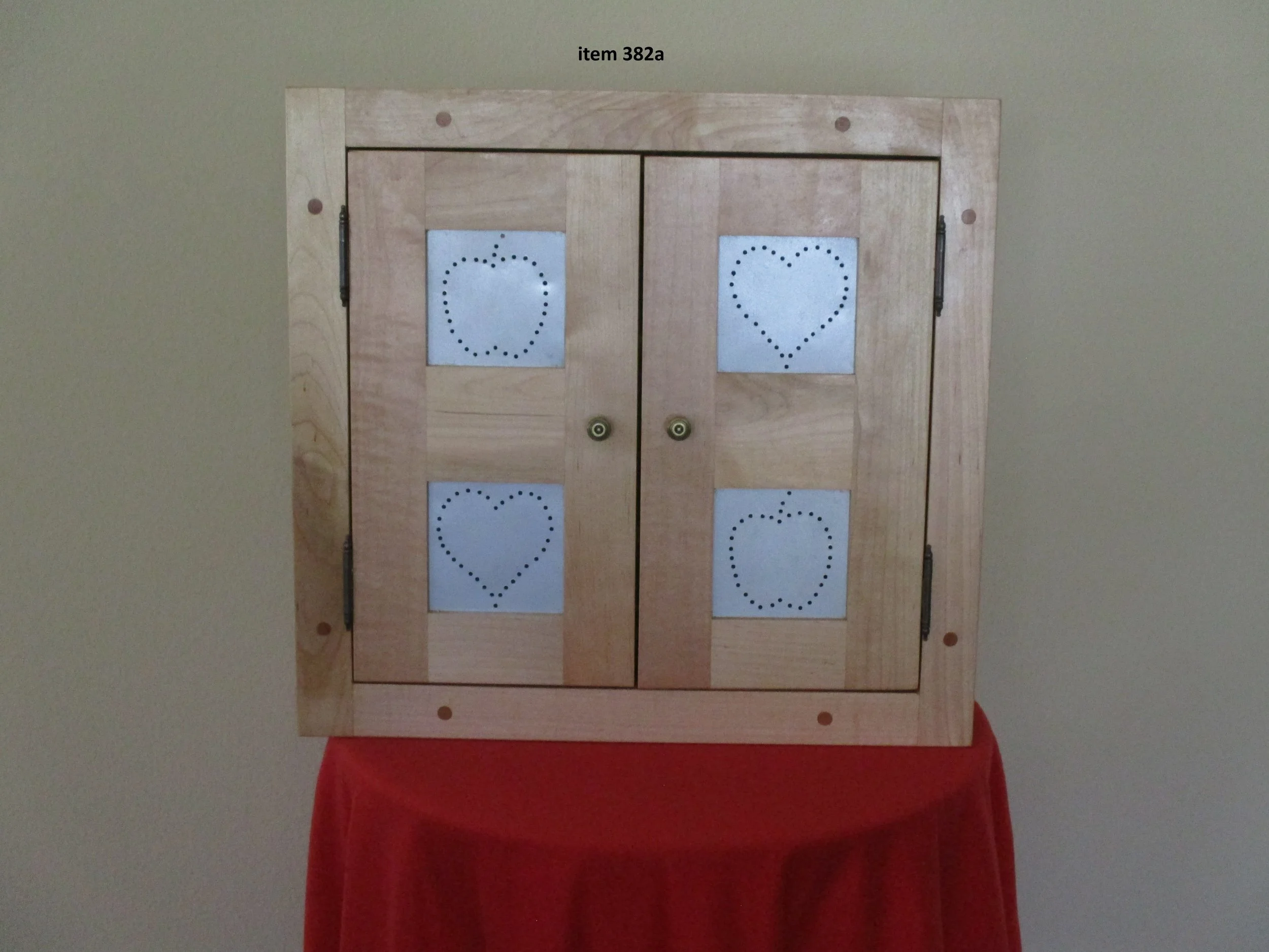 A wooden cabinet with two doors, each featuring two paper sheets with dotted hearts and apples printed on them, mounted on the glass windows. The cabinet is on a red cloth-covered table against a beige wall.