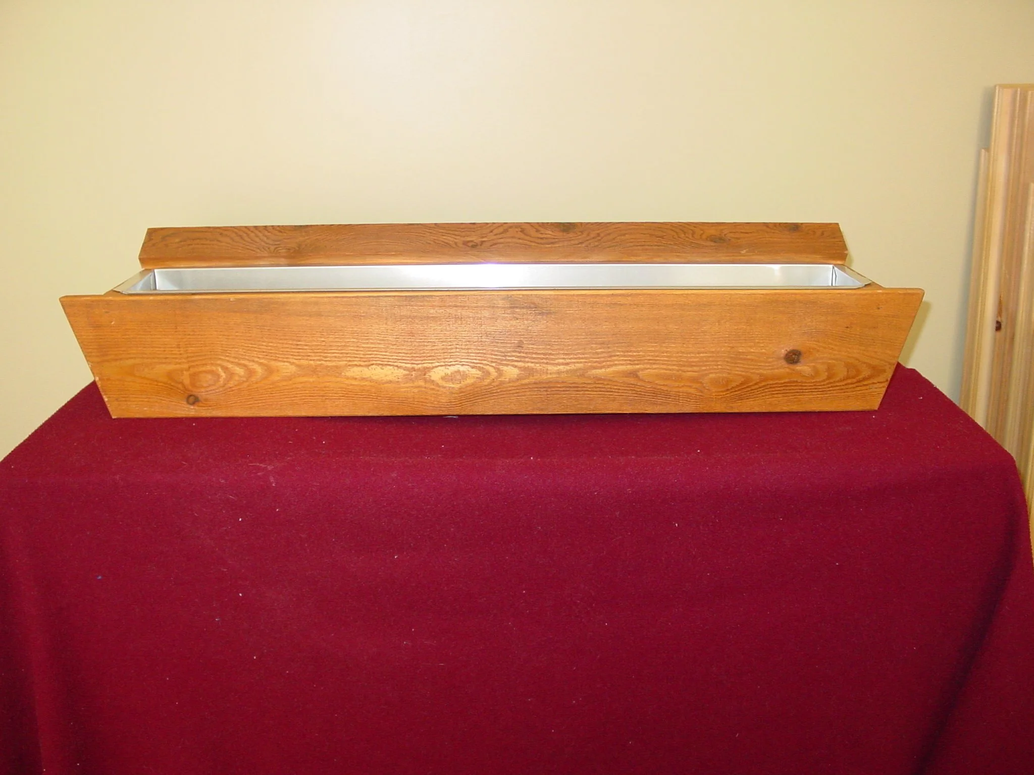 A rectangular wooden planter box with a metal interior, placed on a red tablecloth against a beige wall.