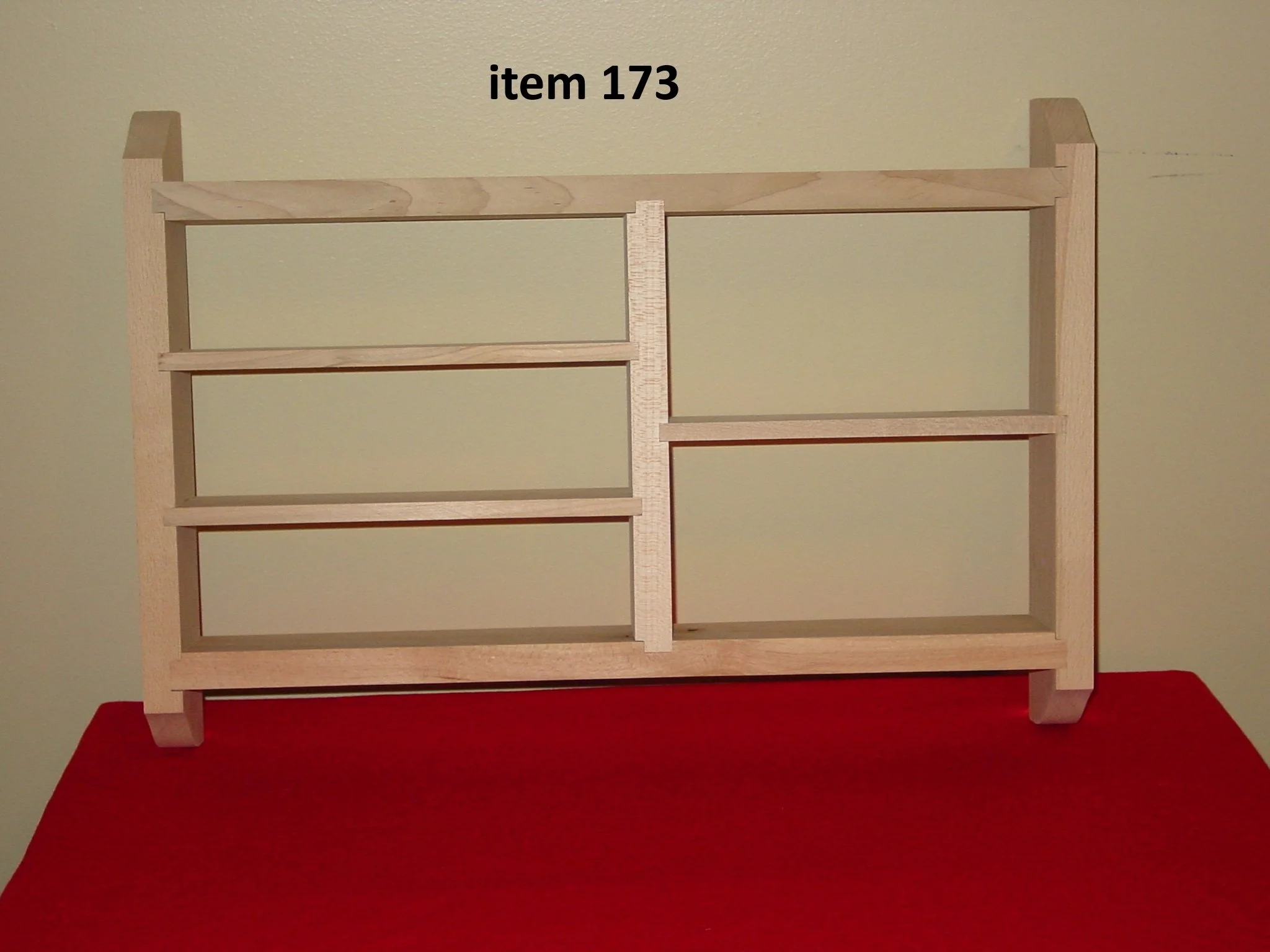 A wooden shelf with multiple compartments, placed on a red surface, against a beige wall, labeled as item 173.