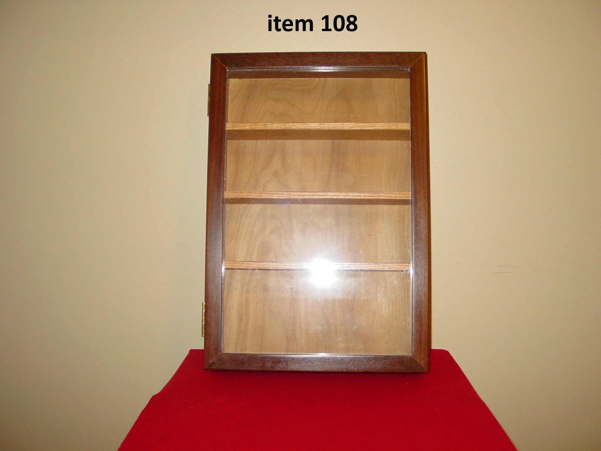 A wooden display case with glass front showing four shelves, placed on a red cloth surface against a beige wall, labeled 'item 108' at the top.