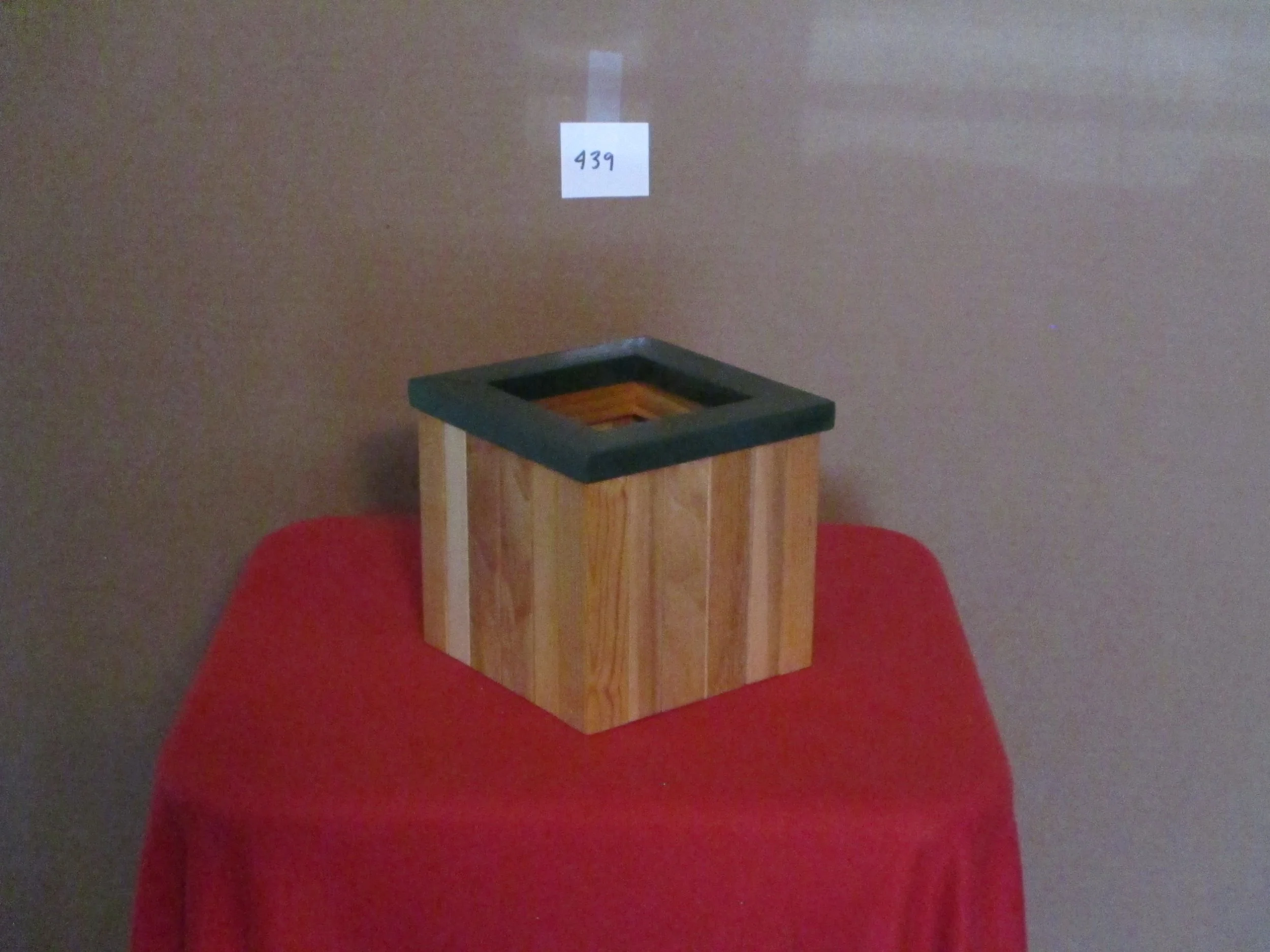 Wooden box with a black frame on top, placed on a red cloth-covered table against a plain wall with a small white sign labeled '439' hanging above it.