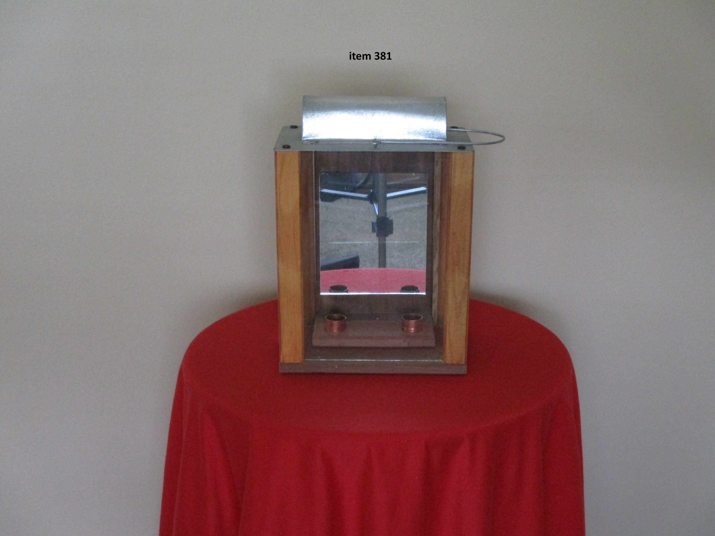 A rectangular display case with wooden sides and a glass front, placed on a red-covered round table against a plain wall. The top has metal covering with a handle. Inside the case are four copper-colored objects.