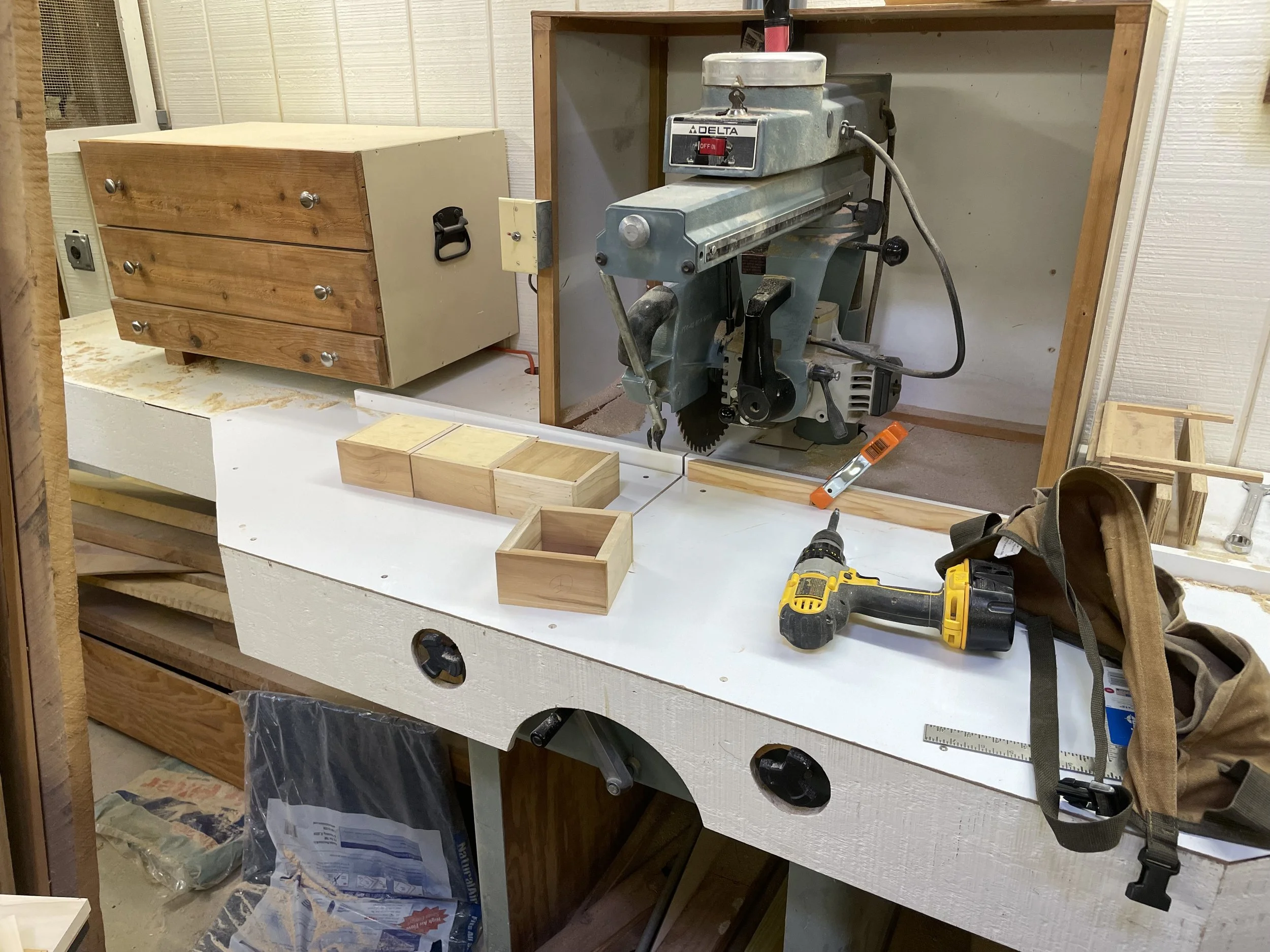 Workshop with a band saw, cordless drill, small wooden boxes, a ruler, and a bag on a white workbench with wooden drawers and a wooden frame in the background.