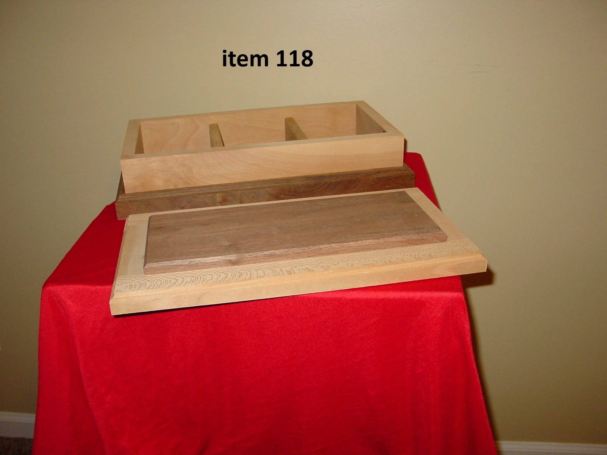 Two wooden cutting boards, one large and one small, placed on a table with a red tablecloth, against a plain beige wall. The larger board has a darker strip of wood in the center, and the smaller board has a raised tray with compartments labeled "item 118".