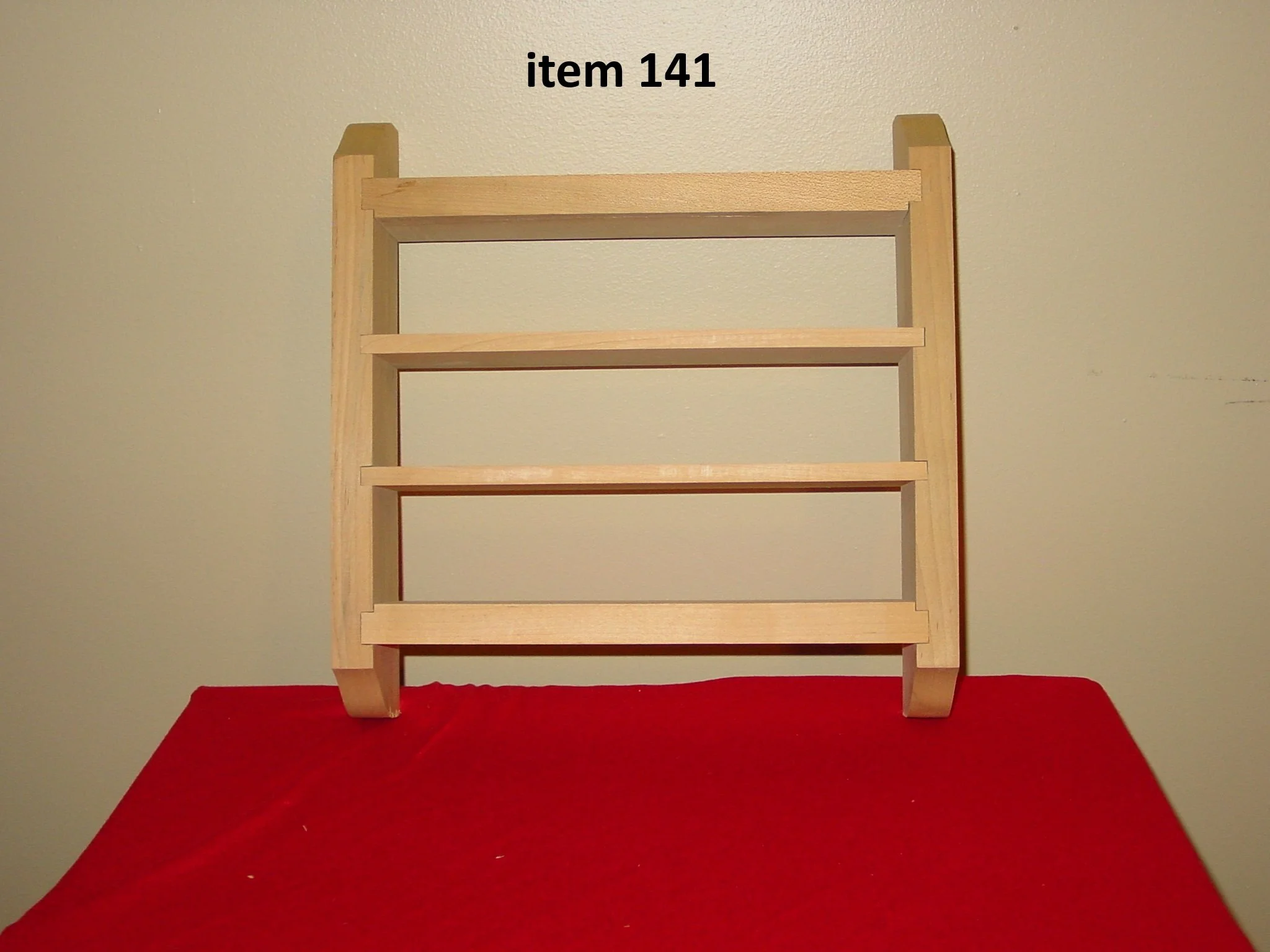A wooden shelf with four horizontal slats, standing against a beige wall, labeled 'item 141' in black text at the top, placed on a red surface.