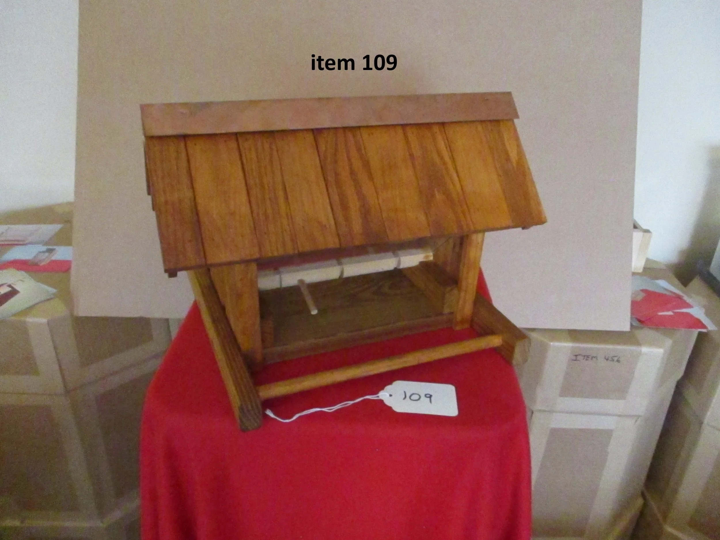 Small wooden house model with labeled item 109, placed on a red cloth and surrounded by boxes.