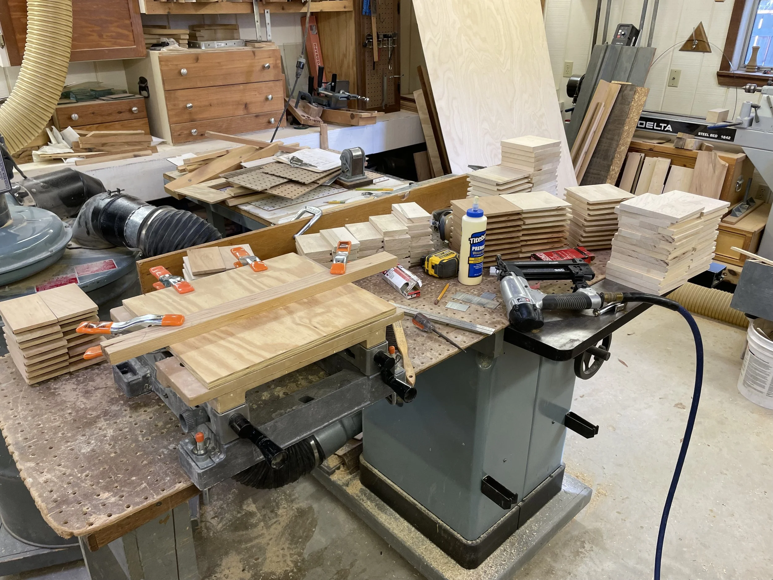 Woodworking workshop with stacks of wood pieces, clamps, power tools, glue, and various woodworking supplies on a workbench.