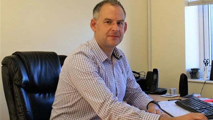 Gareth Botterill sitting at a desk in an office, wearing a checkered shirt, with a computer keyboard, phone, and papers on the desk.