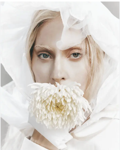 A person with blonde hair holding a white flower in front of their mouth, surrounded by white fabric.