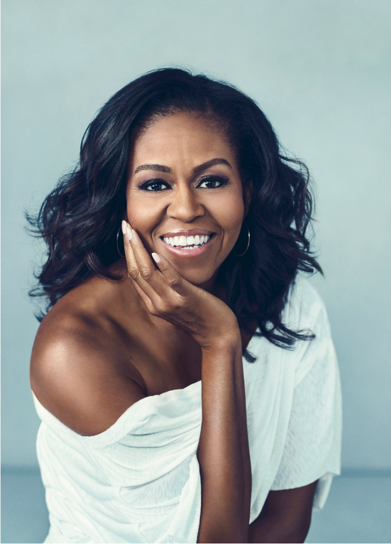 A woman with dark, wavy hair smiling and resting her chin on her hand, wearing a white off-the-shoulder top and hoop earrings.