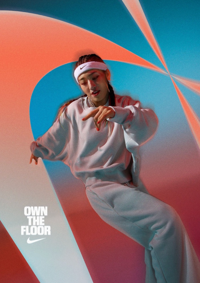 A woman in a white Nike outfit and headband dances in front of a colorful abstract background with geometric shapes and lighting effects, with the words "OWN THE FLOOR" and Nike logo in the bottom left corner.