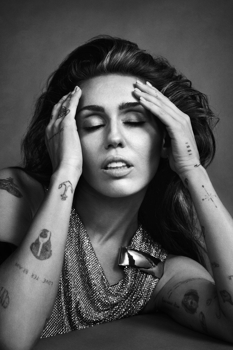 Black and white portrait of a woman with long, wavy hair, holding her head with both hands, eyes closed, wearing a sparkly top and jewelry, with tattoos visible on her arms.