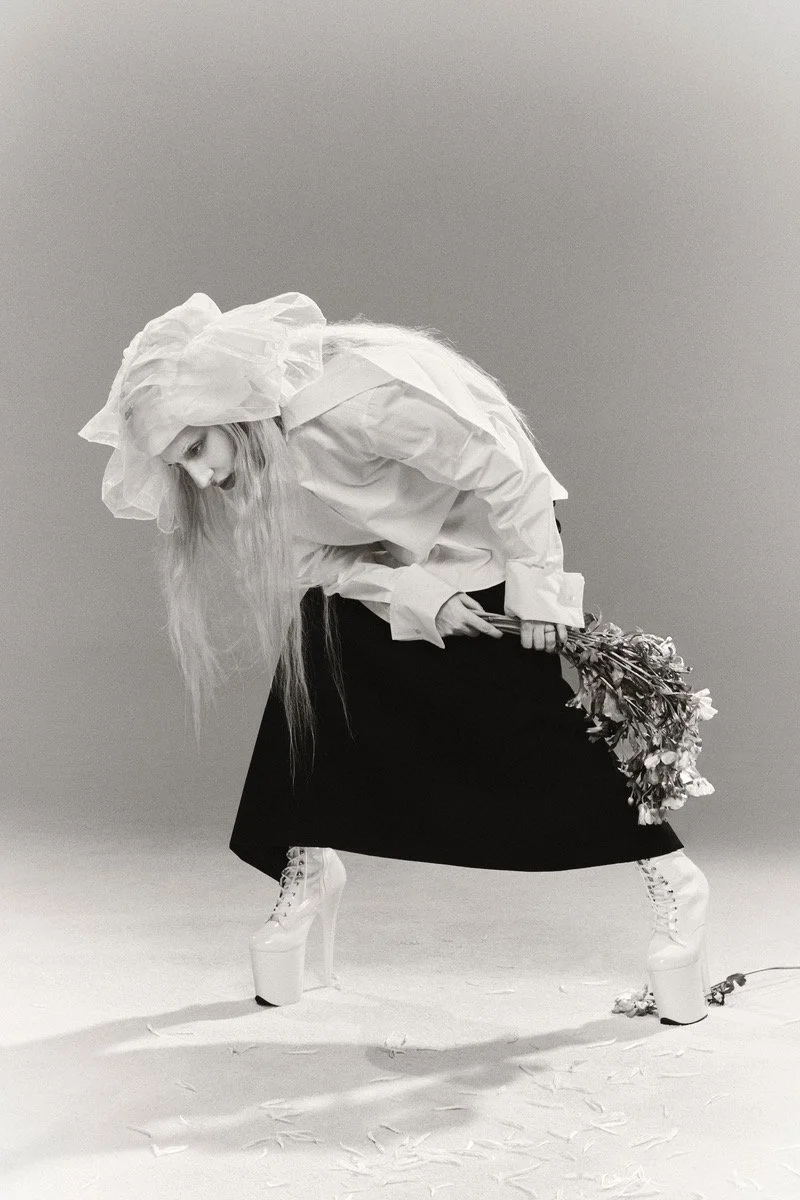 A woman with long hair dressed in a white shirt and a black skirt, wearing high platform heels, holding a bunch of flowers behind her back in a studio with a plain background.