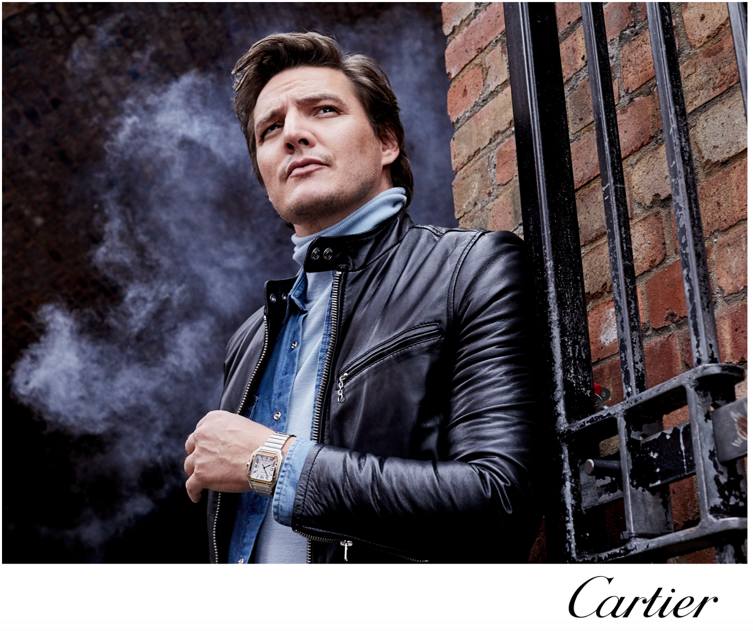 A man in a leather jacket standing next to a brick wall with metal bars, looking thoughtfully into the distance.