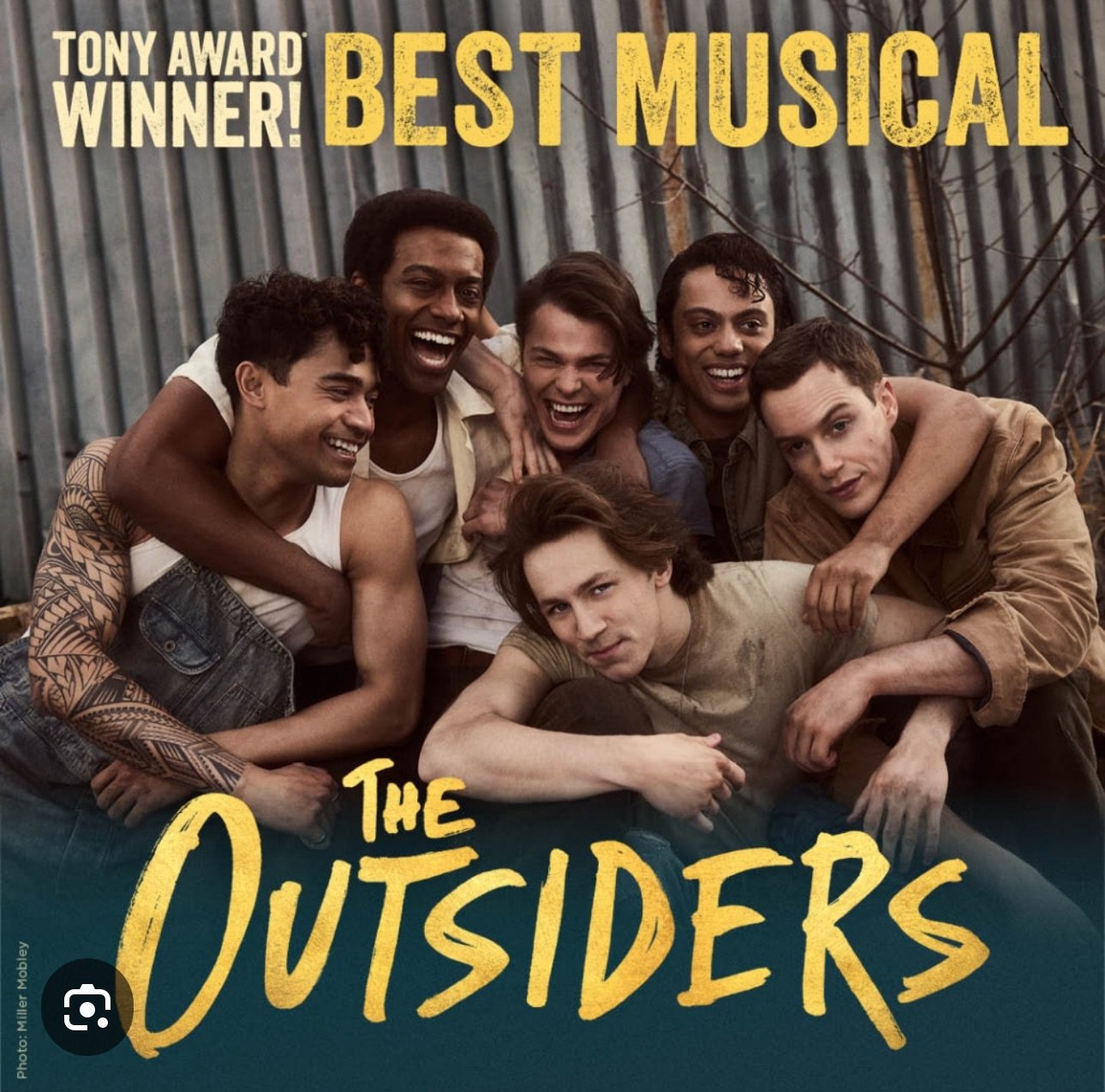 Group of seven young men huddled together outdoors in front of a metal background, smiling and showing camaraderie. Text overlay reads "Tony Award Winner! Best Musical. The Outsiders."