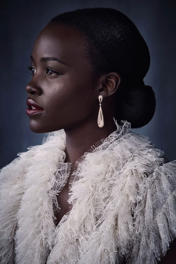 A woman with dark skin and elegant makeup, wearing a large, white, textured lace garment with ruffles, and long, dangling earrings, posing against a dark background.