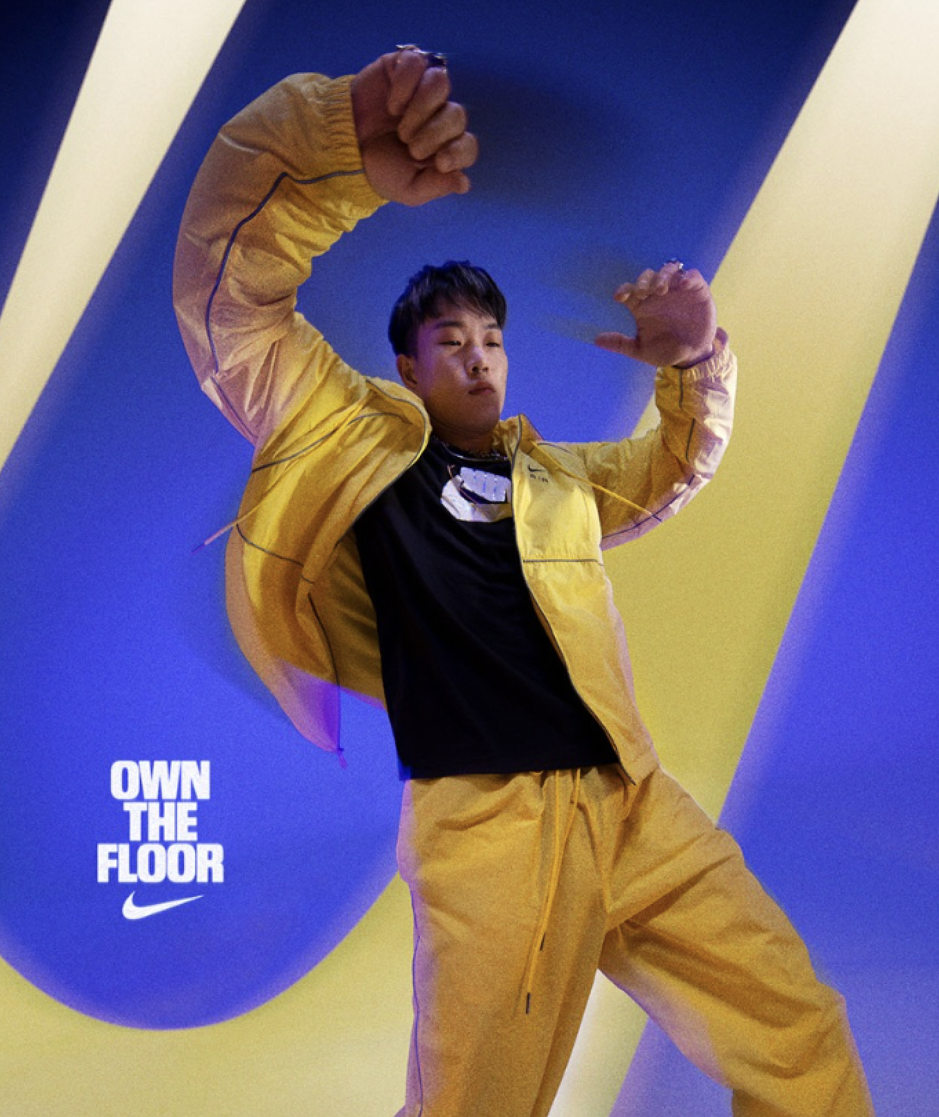 A young man wearing a yellow tracksuit and black t-shirt posing in front of a blue background with yellow accents. The text 'OWN THE FLOOR' and the Nike swoosh logo are visible in the lower left corner.