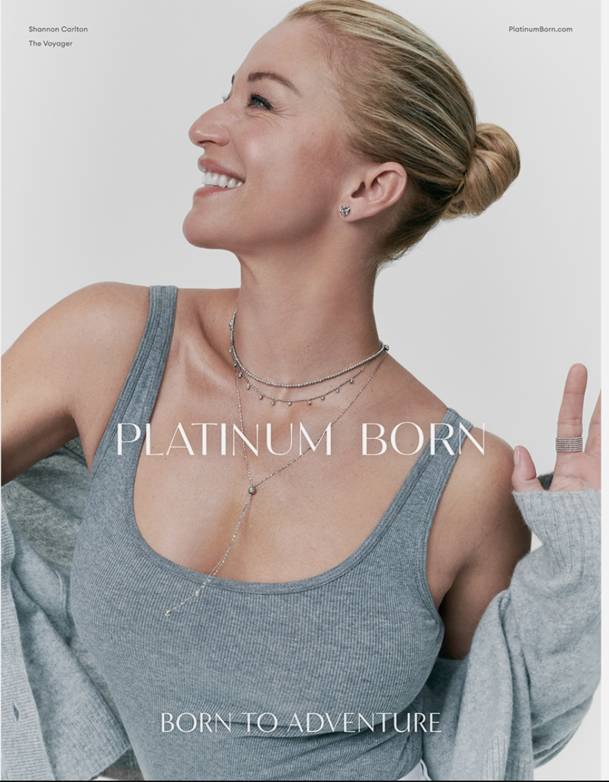 A woman smiling and making a peace sign with her right hand. She has blonde hair tied back in a bun and is wearing jewelry including earrings, layered necklaces, and rings. She is dressed in a gray tank top and a gray cardigan, with a plain light bac