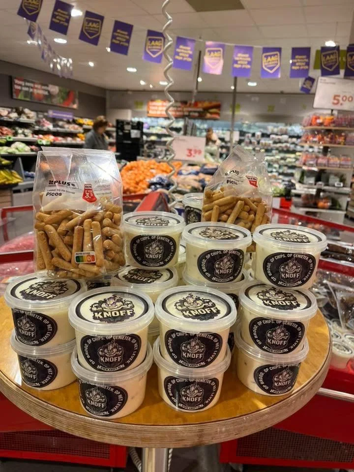 Verschillende bakjes met knoflooksaus en zakjes met knapperige snacksticks op een toonbank in een supermarkt.