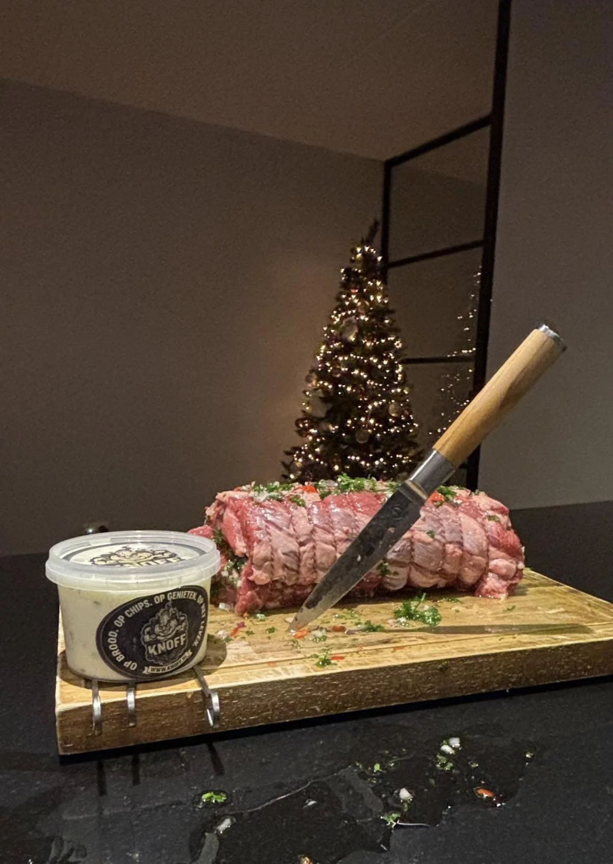 Vlees op een snijplank met een mes, een bakje kruiden of saus en een kerstboom op de achtergrond.