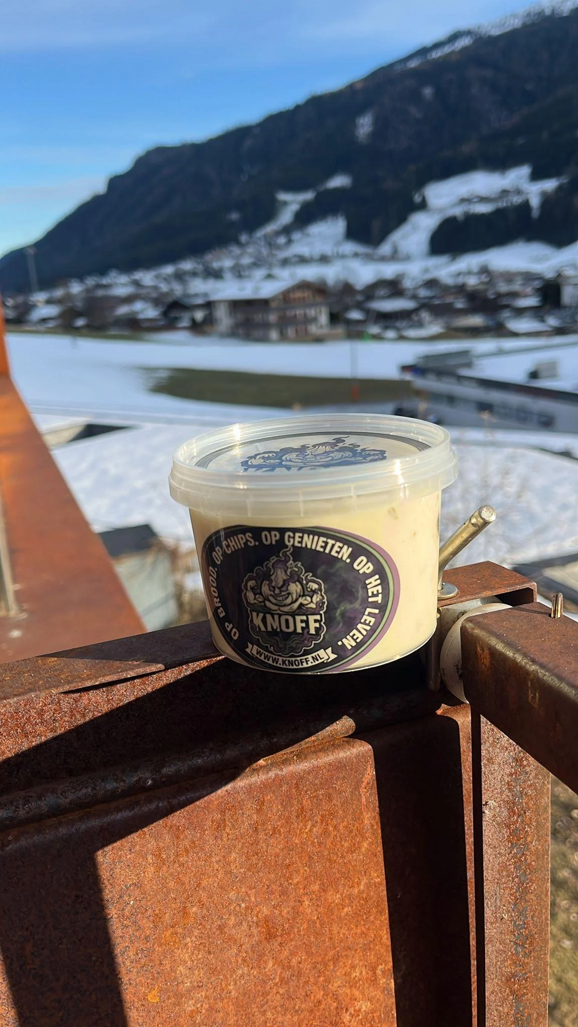 Bodempot met knof-lookdip op een metaalriem tegen een bergachtig landschap met sneeuw en een dorp.