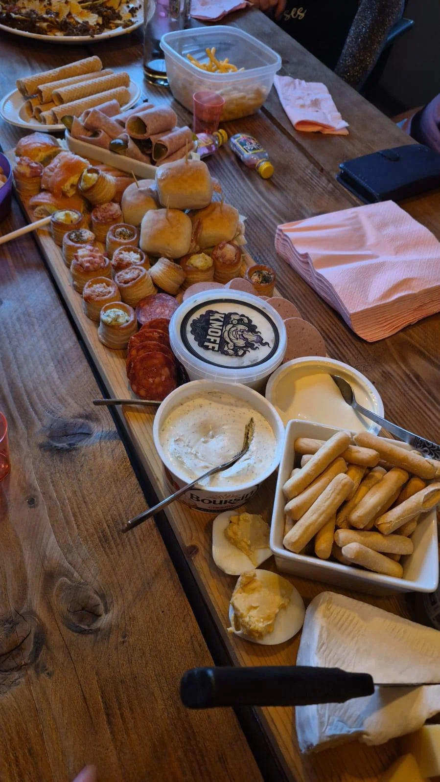 Assortiment van snacks en dips op een houten tafel, inclusief mini sandwiches, rolletjes, worstjes, kaas, chips, sauzen, en stokbroodjes.