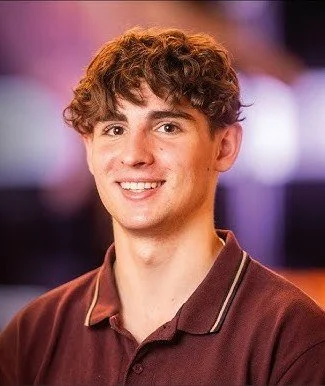 Junger Mann mit braunen, lockigen Haaren, trägt ein braunes Polohemd, lächelt vor bunter, unscharfer Hintergrund.