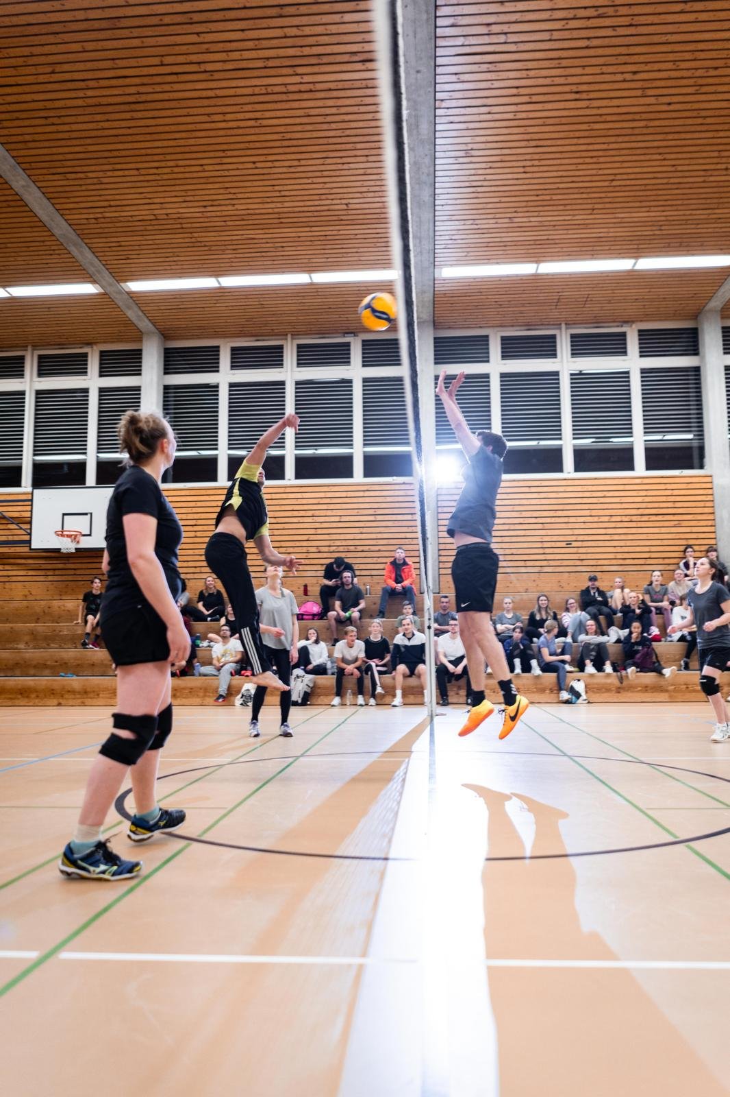 Zwei Volleyballspieler springen und schlagen den Ball über das Netz, während ein weiteres Teammitglied zuschaut, in einer Turnhalle mit Zuschauern auf den Bänken.