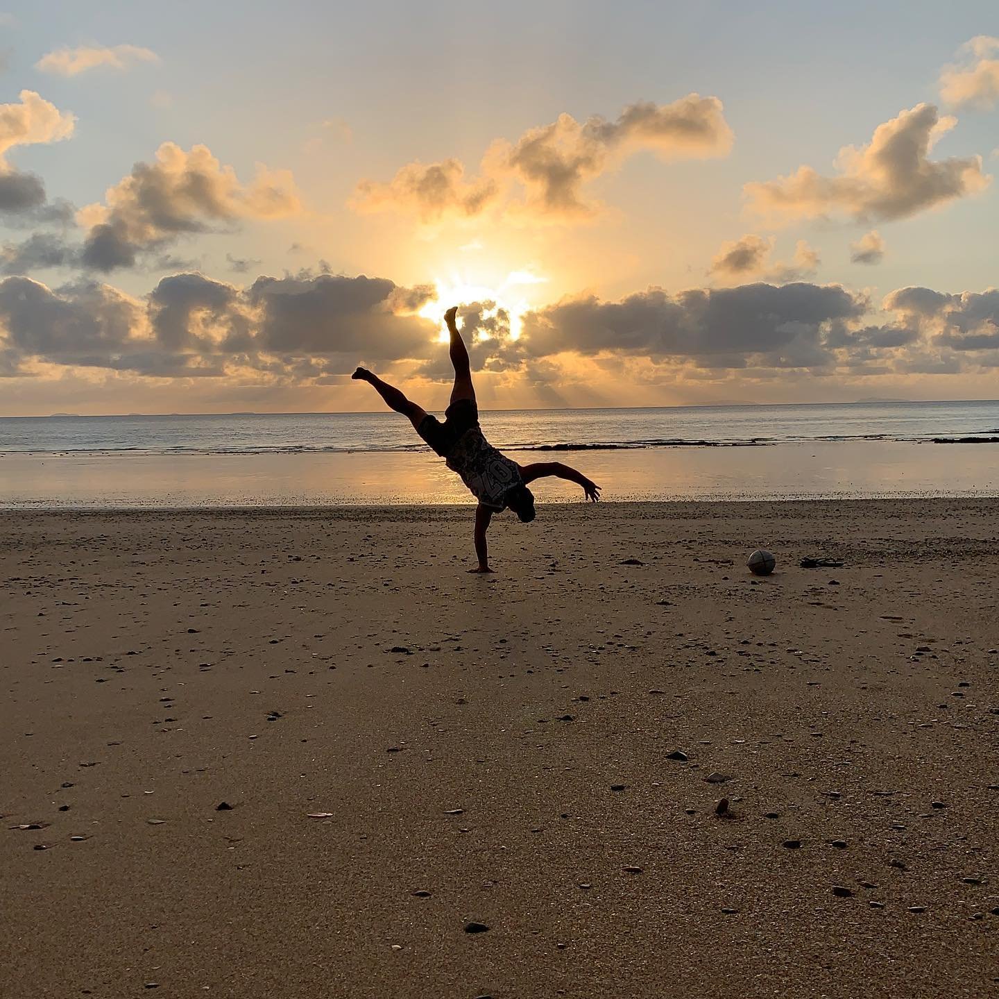 Person performing a handstand on a sandy beach during sunset, with clouds in the sky and an ocean in the background.