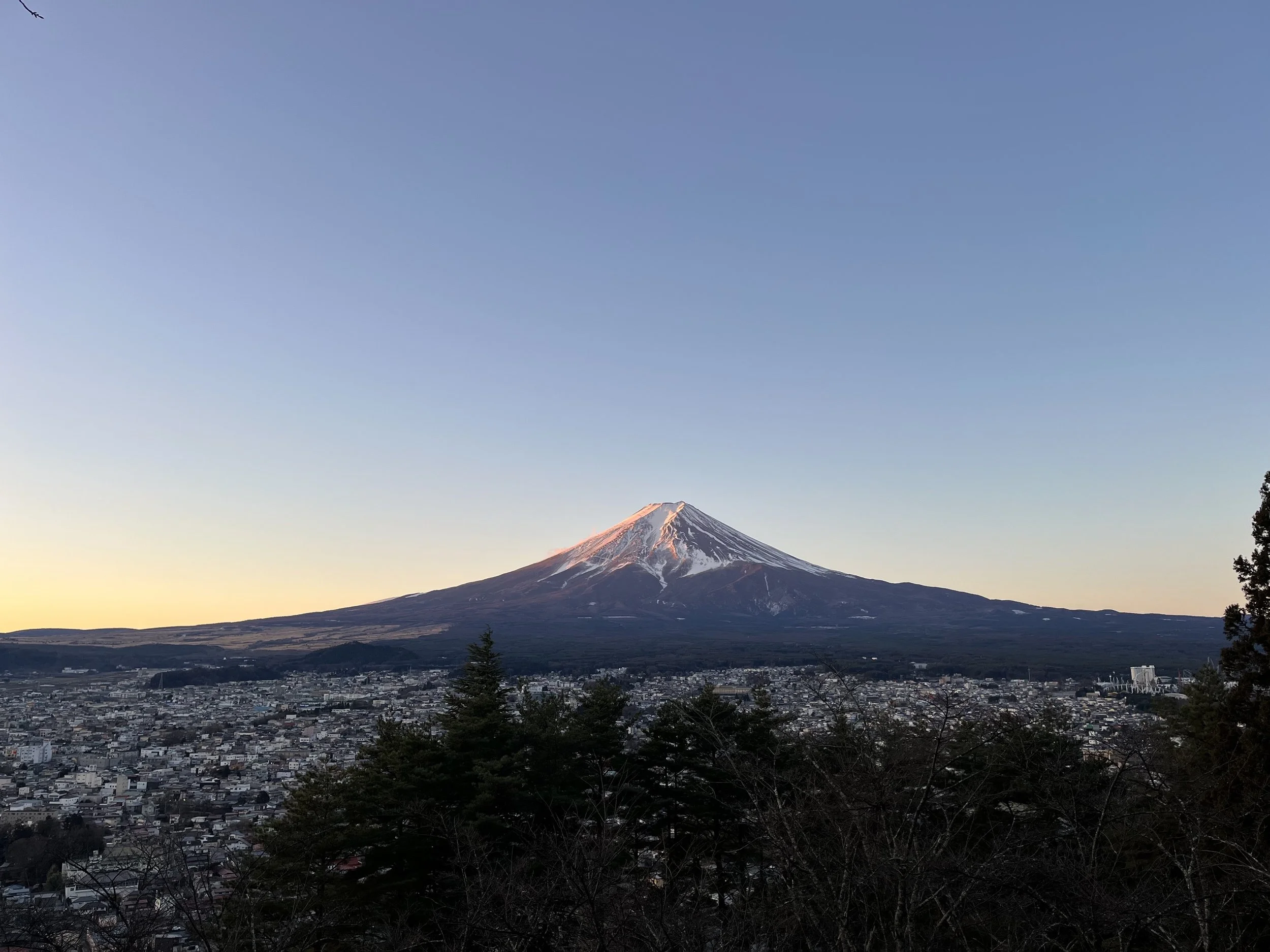 Snow-capped Mount Fuji during sunset, with a city in the foreground and trees at the bottom.