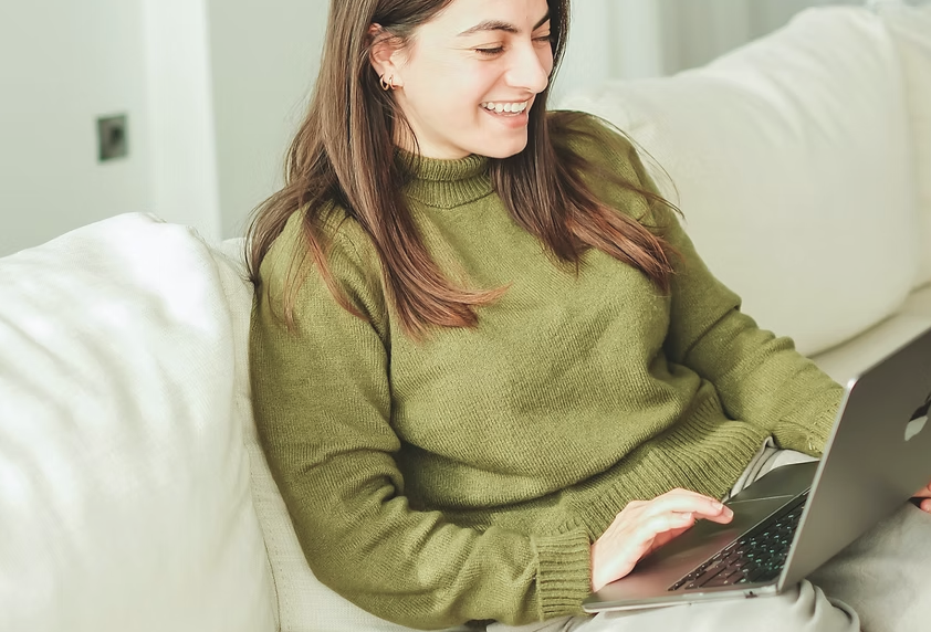 A young woman with brown hair, wearing a green sweater, sitting on a white couch and smiling while using a laptop.