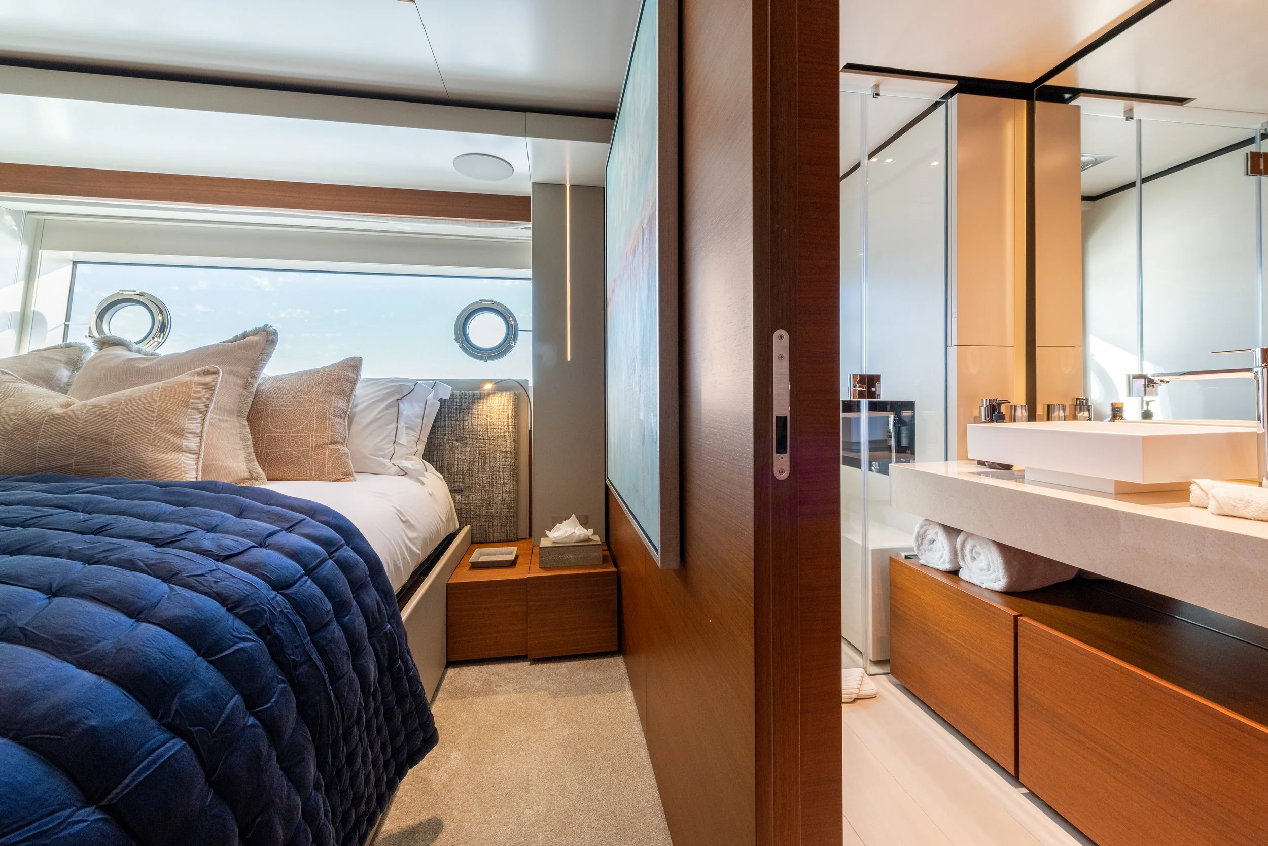 Modern bedroom with a bed, pillows, side table, and a large window with portholes, visible bathroom with sink, towels, and shower area.