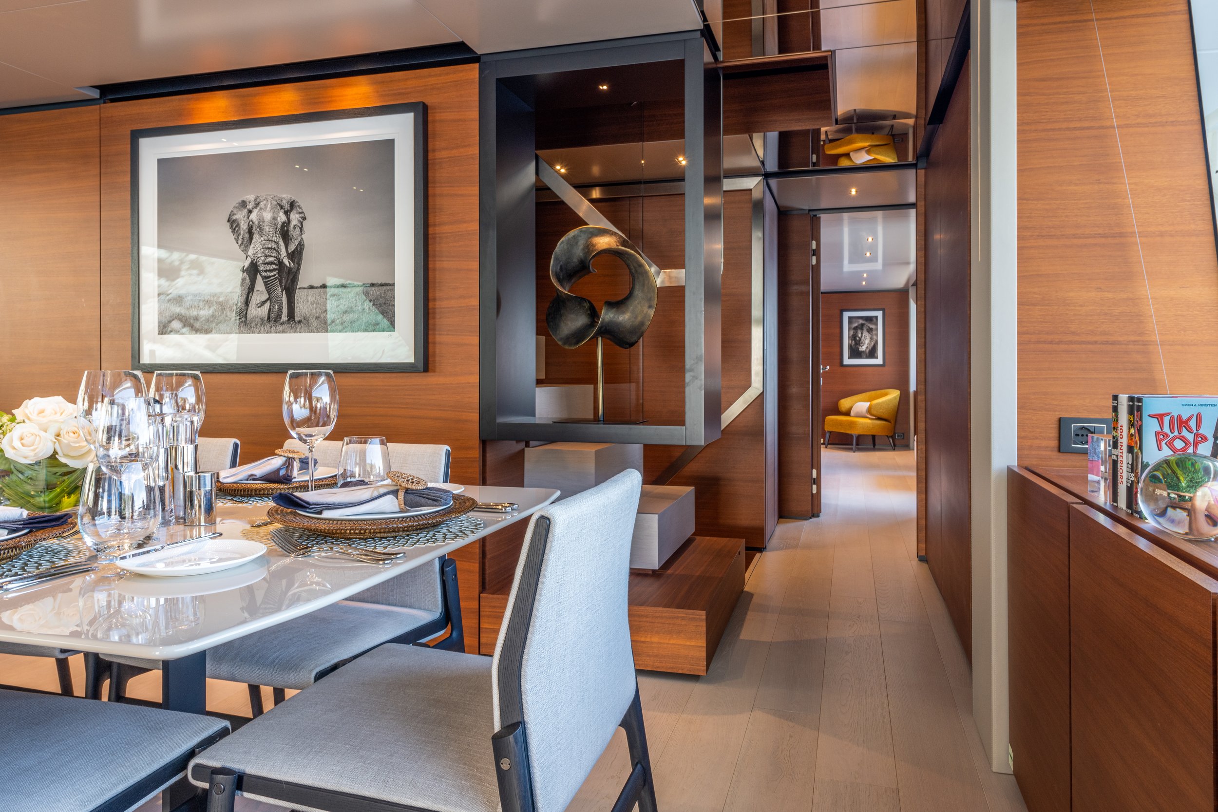 Modern dining room with a wooden wall, a table set with wine glasses and napkins, a framed black and white elephant photograph on the wall, and a decorative sculpture on a shelf. A hallway with chairs and artwork is visible in the background.