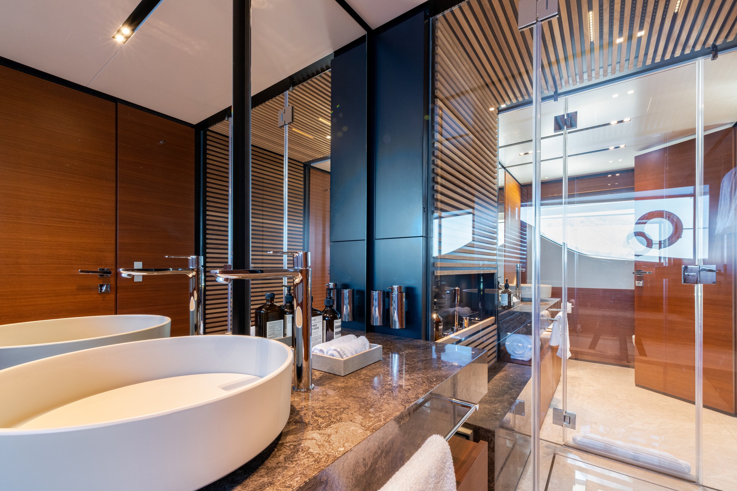 Modern hotel bathroom with a bathtub on a marble countertop, glass shower door, wooden wall panels, and large windows letting in natural light.