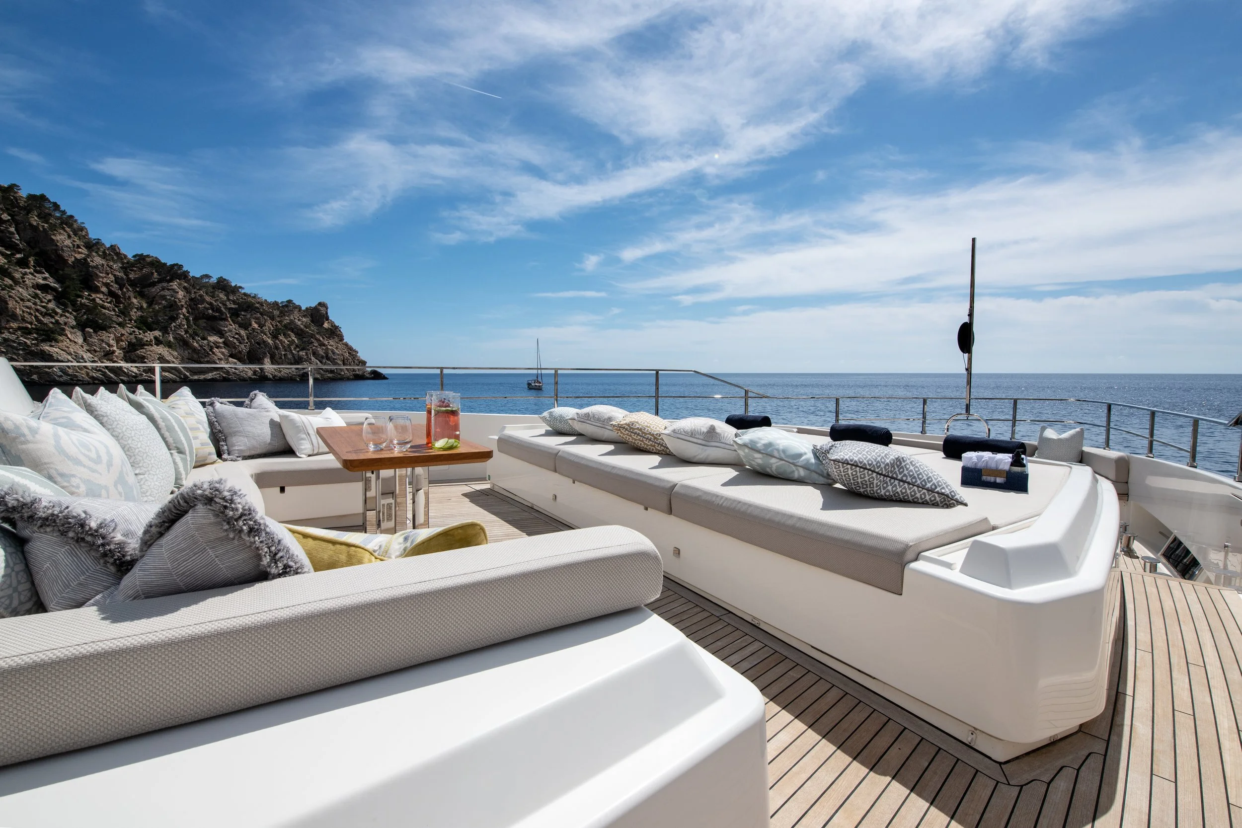 Luxury yacht deck with cushioned seating and pillows, overlooking the ocean and a rocky coastline under a blue sky.