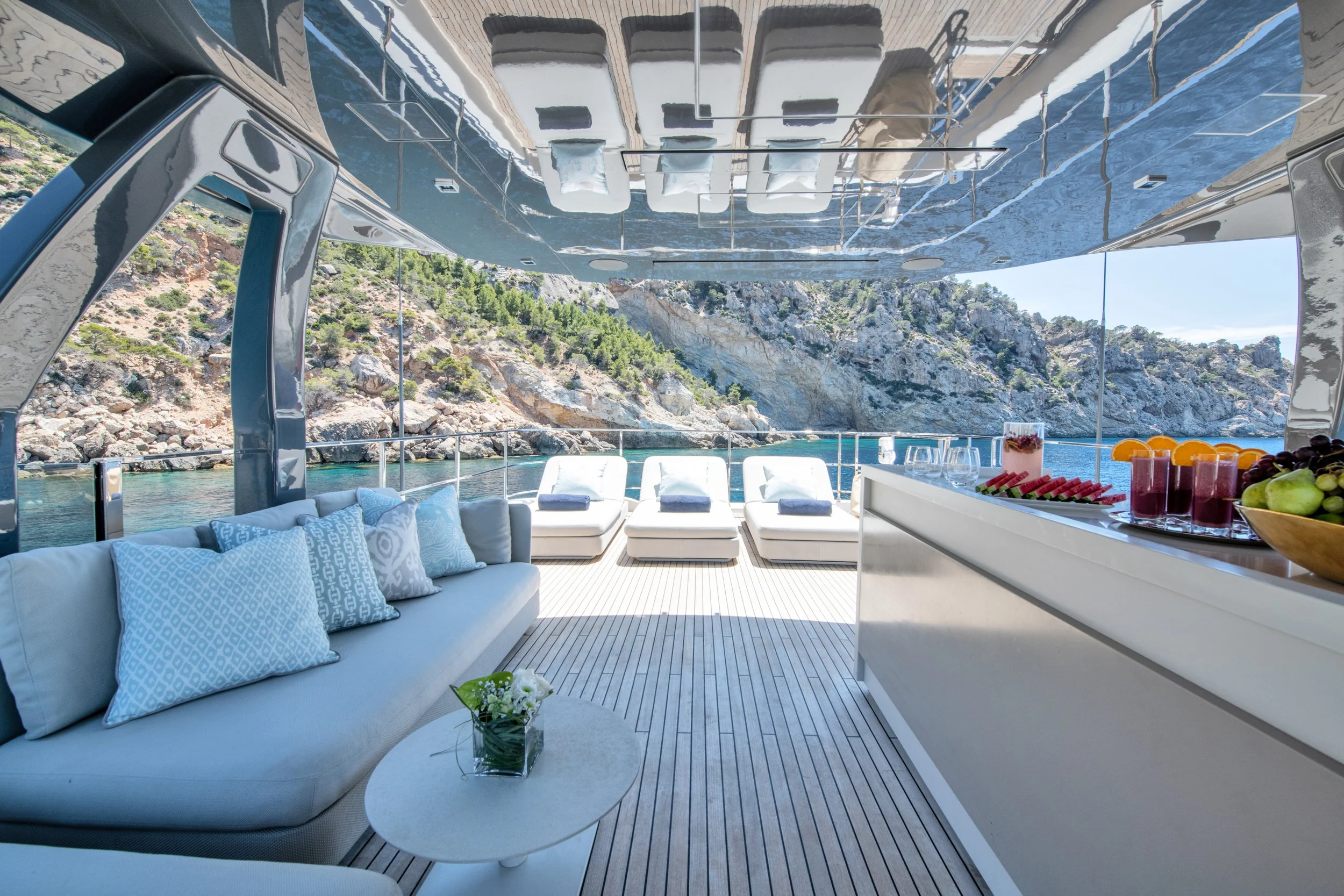 Outdoor yacht deck with white cushioned lounge chairs overlooking rocky coastline and water, modern white seating area with pillows, bar with fresh fruit and drinks, and a small table with flowers, under a shiny metallic ceiling.