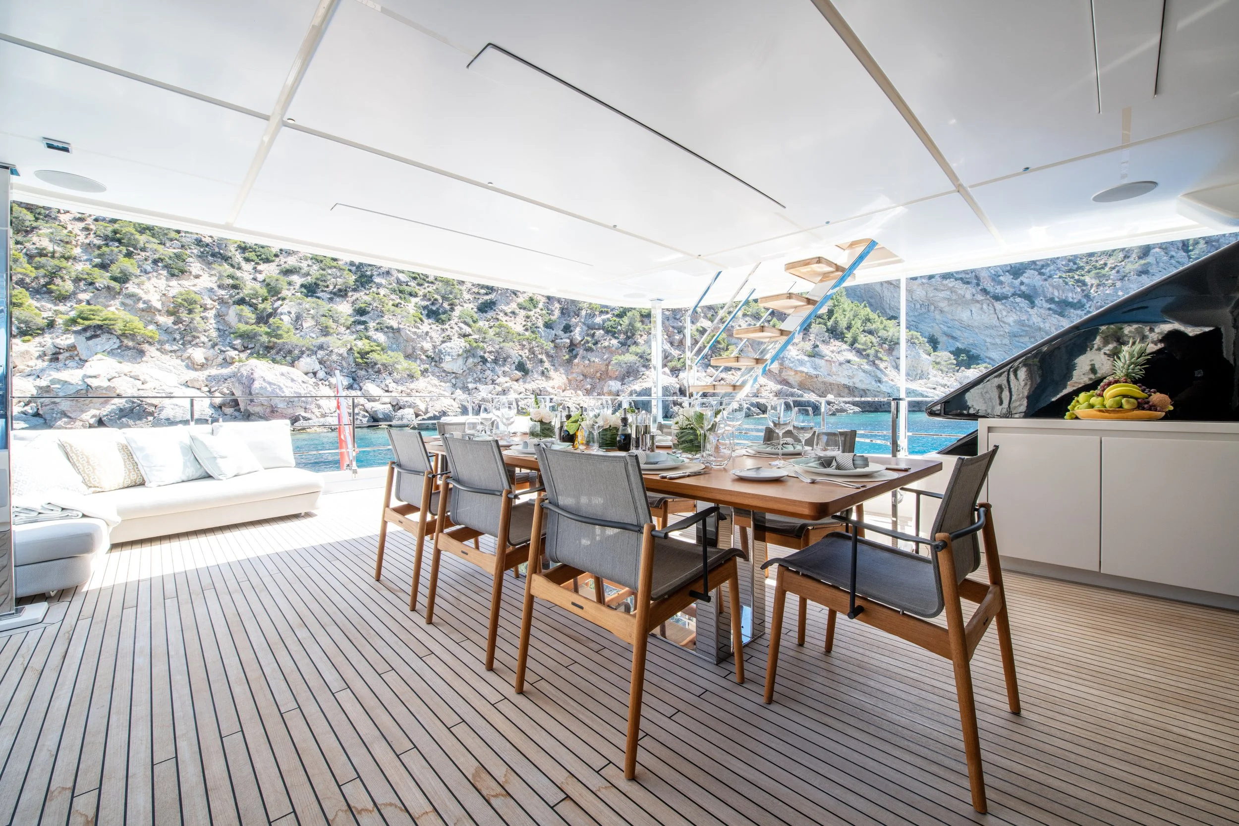 Elegant outdoor dining area on a yacht with a wooden table set for a meal, surrounded by chairs, overlooking a rocky shoreline with water and hills.