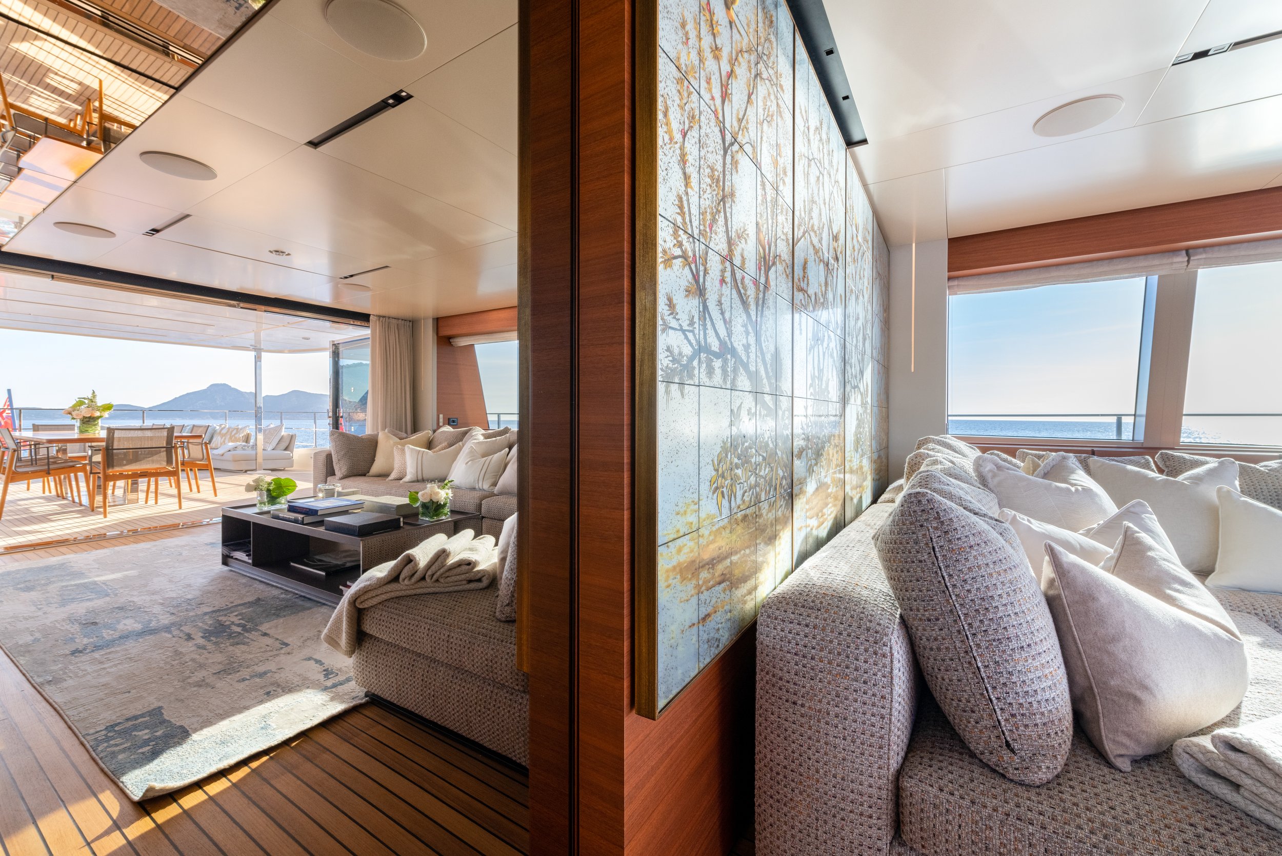 Living room with large windows showing an ocean view, beige couches with white pillows, a painting or mural on a wooden panel, and a dining area on a deck with outdoor furniture.
