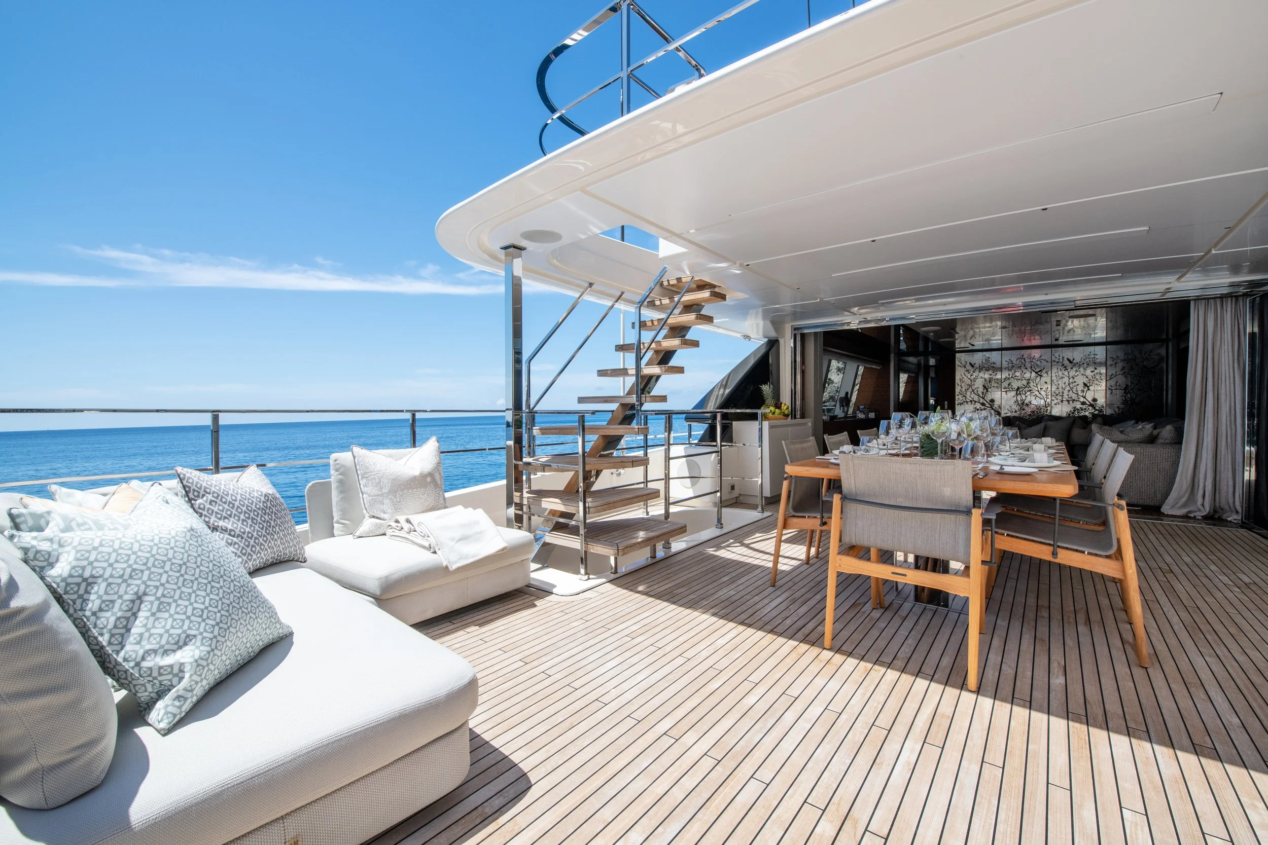 Outdoor deck on a yacht with a seating area, dining table set for a meal, and a staircase leading to the upper deck, overlooking the ocean under a clear blue sky.