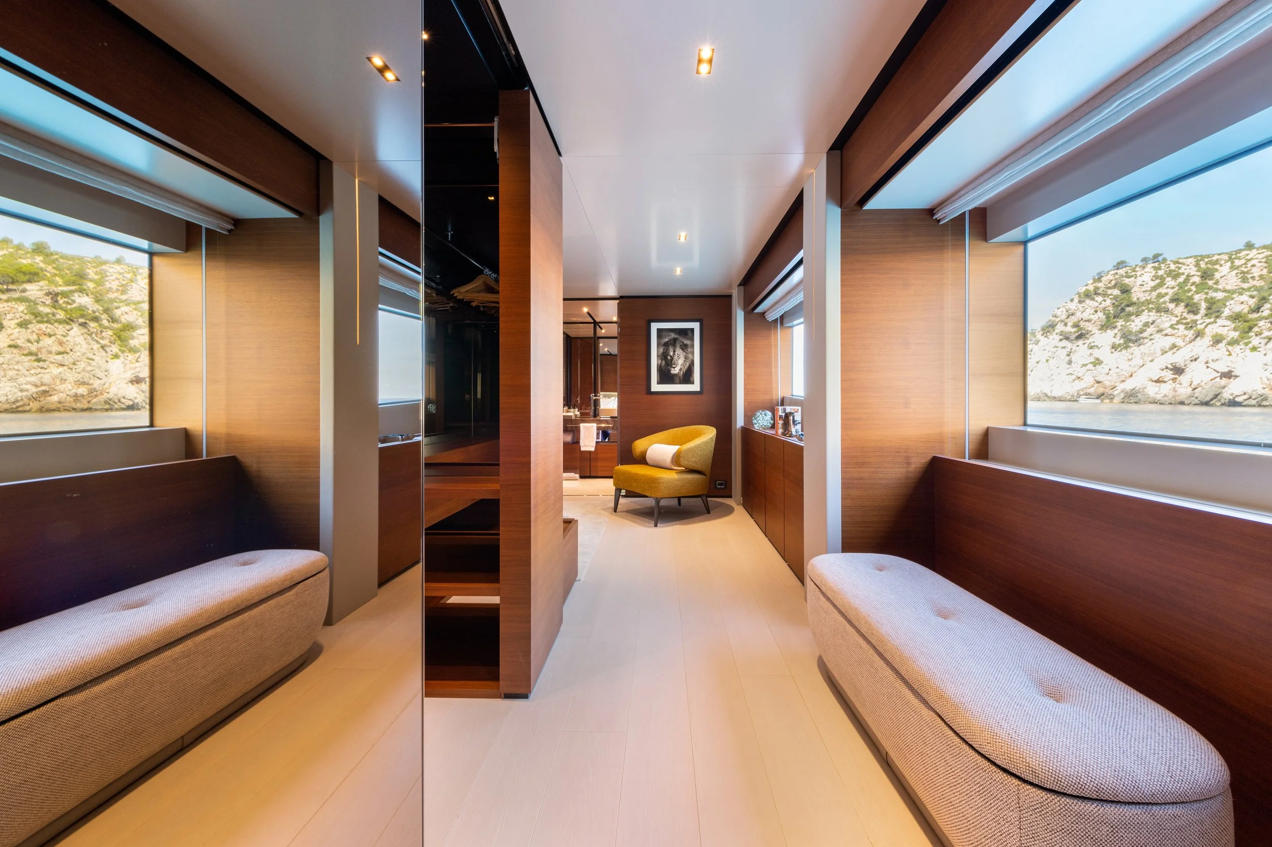 A modern interior room with large windows showing a view of water and rocky hills, two beige cushioned benches, a yellow armchair with a pillow, and a framed black and white image of a lion.