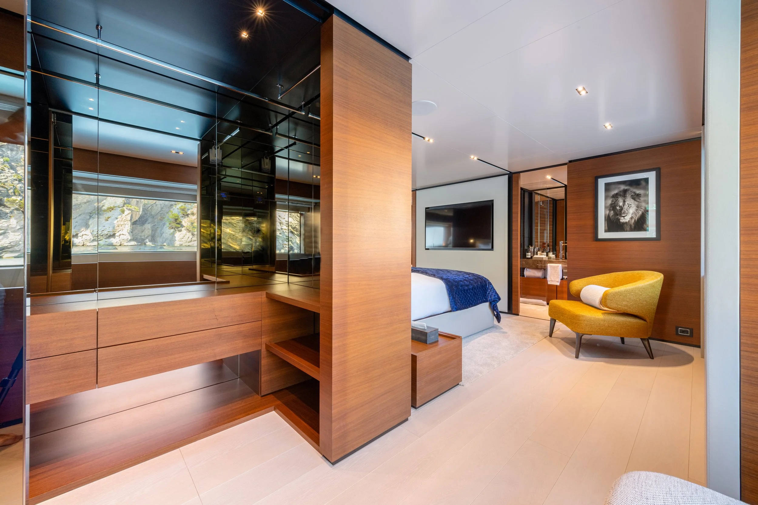 Modern hotel room with wood-paneled walls, a bed with a blue throw blanket, a yellow armchair, a large wall-mounted TV, and a bathroom vanity area in the background.