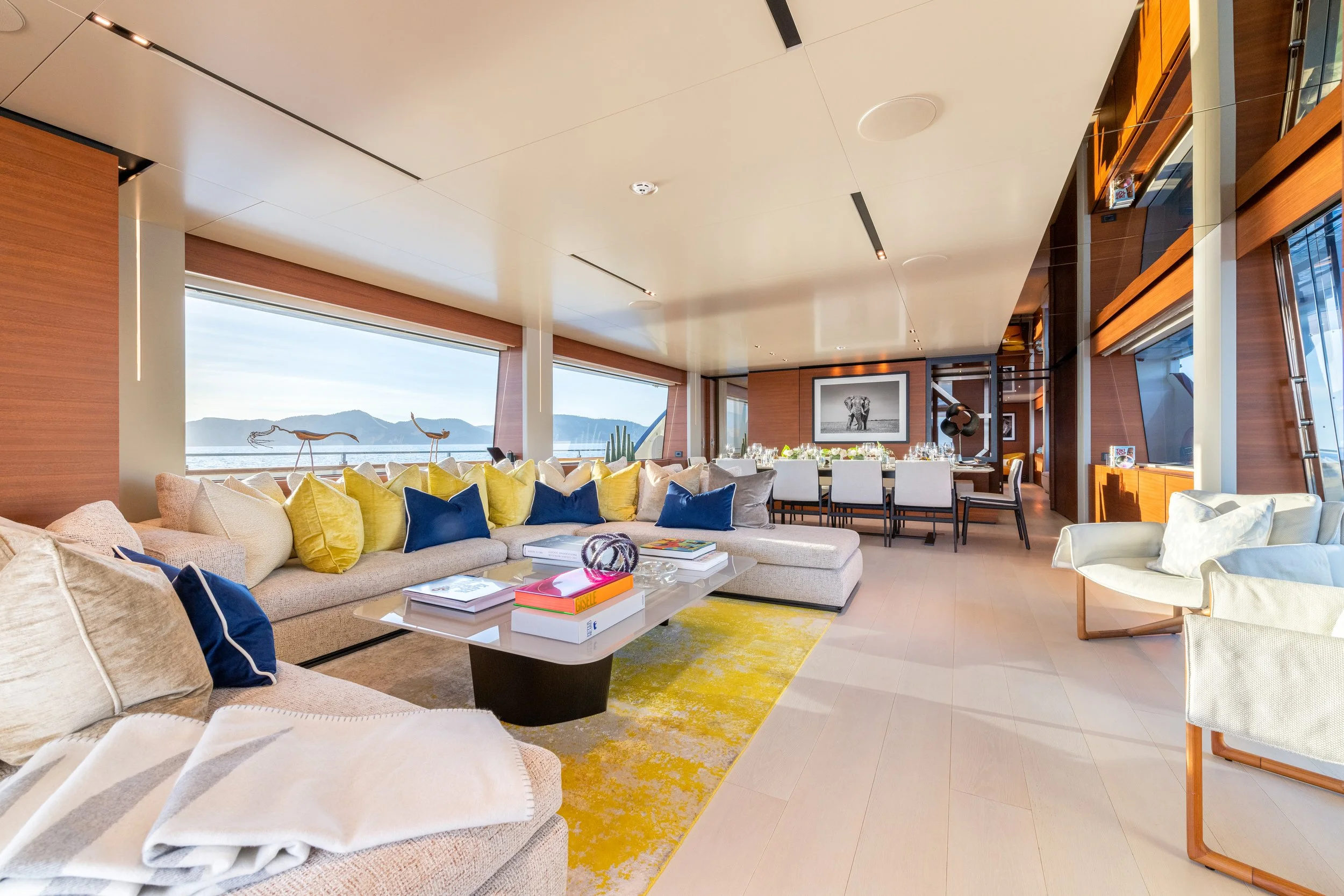 Modern living room with large windows showing mountain views, a beige sectional sofa with yellow, blue, and beige cushions, a white armchair, a white coffee table with books and decorative items, and a yellow rug. There's a dining table with white ch