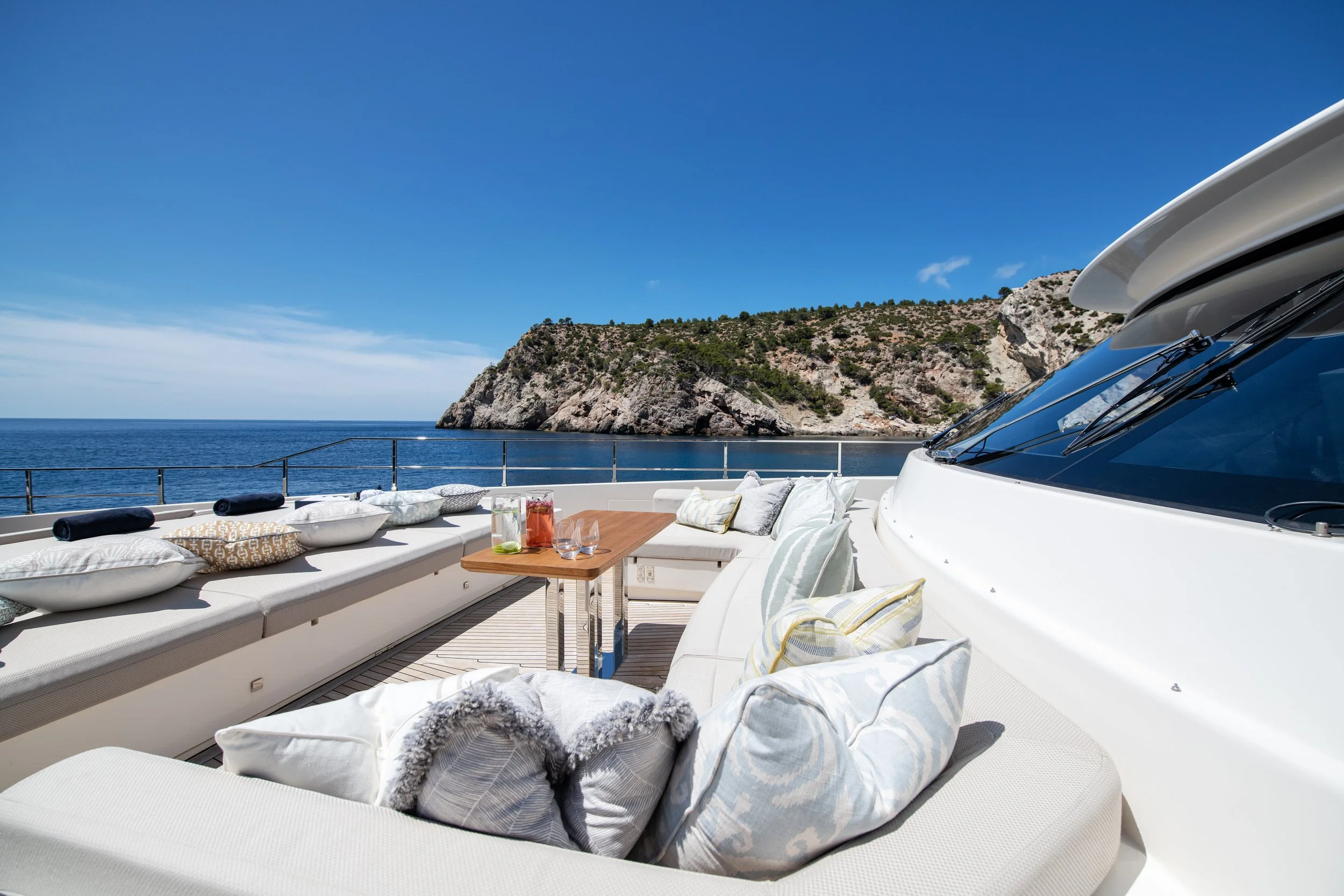 Luxury yacht deck with pillows and a table overlooking the ocean and rocky coastline under a blue sky.